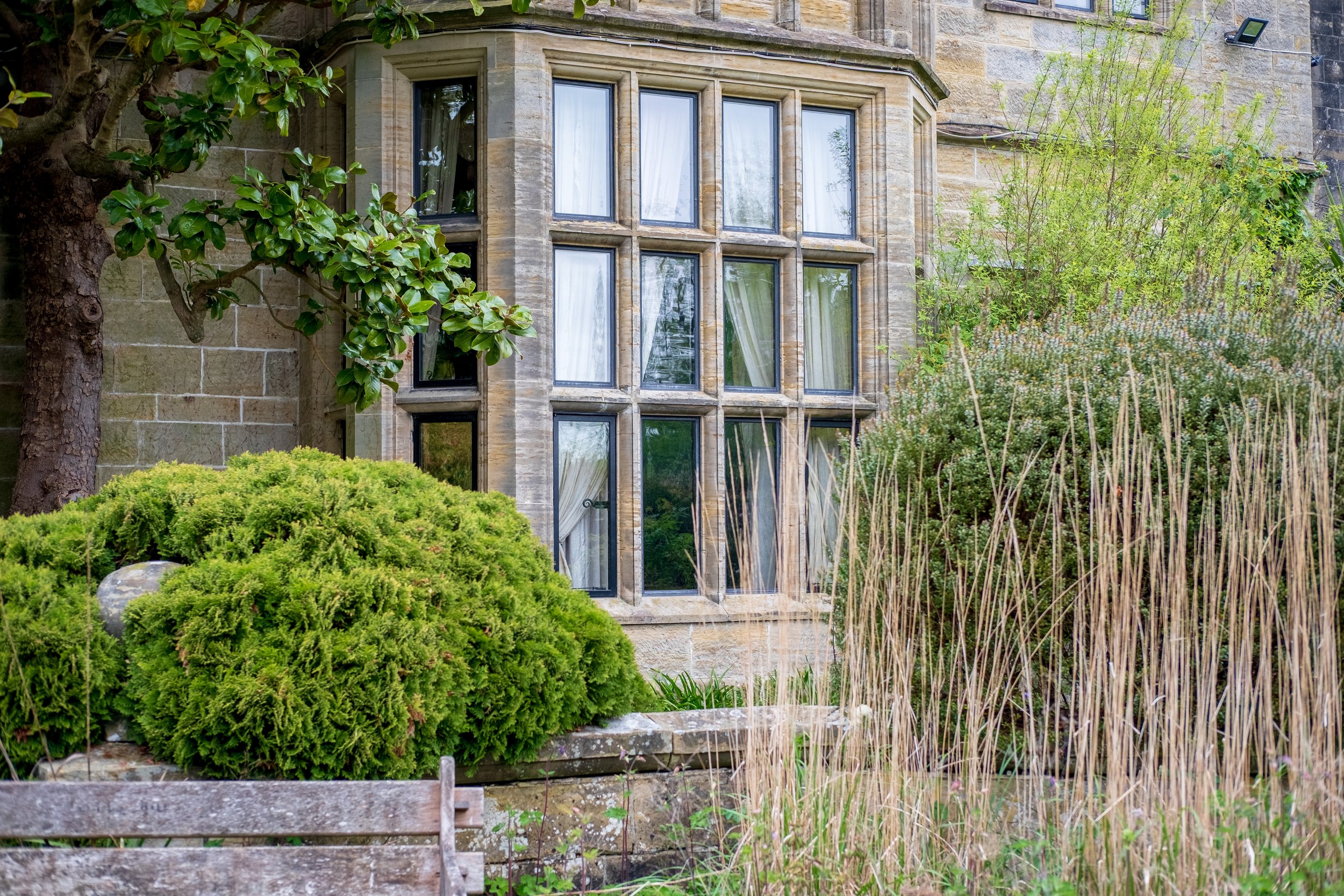 A stone building with large bay windows surrounded by lush green plants and trees.