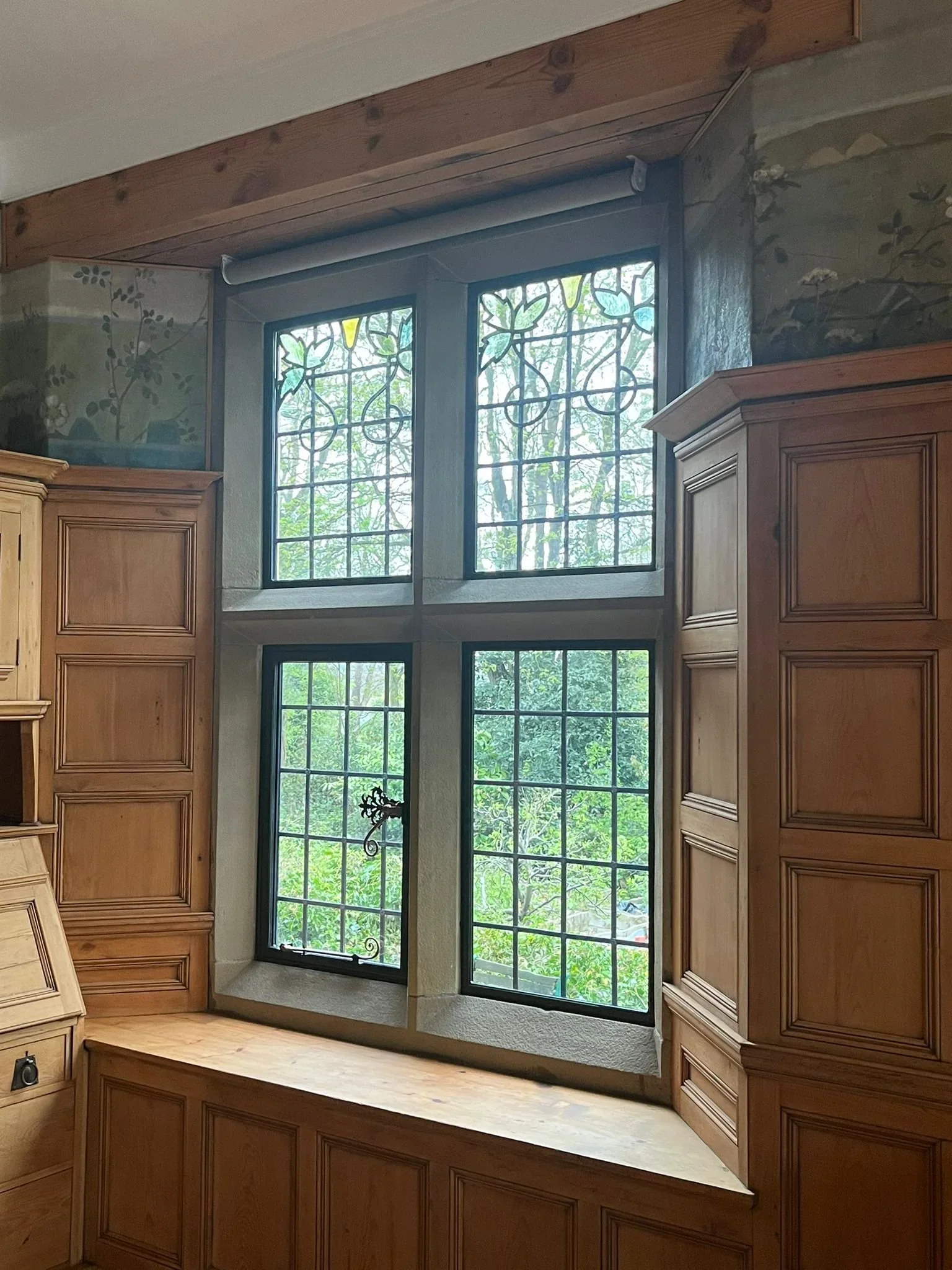 A set of four vertical stained-glass windows with decorative black grid patterns, surrounded by wooden trim, with trees visible outside.