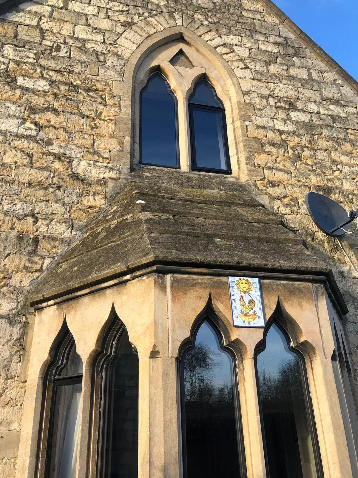 Close-up of a stone church building with Gothic-style arched windows, a small decorative window at the top, a satellite dish on the right, and a colorful rooster and sun emblem above the large stained glass windows.