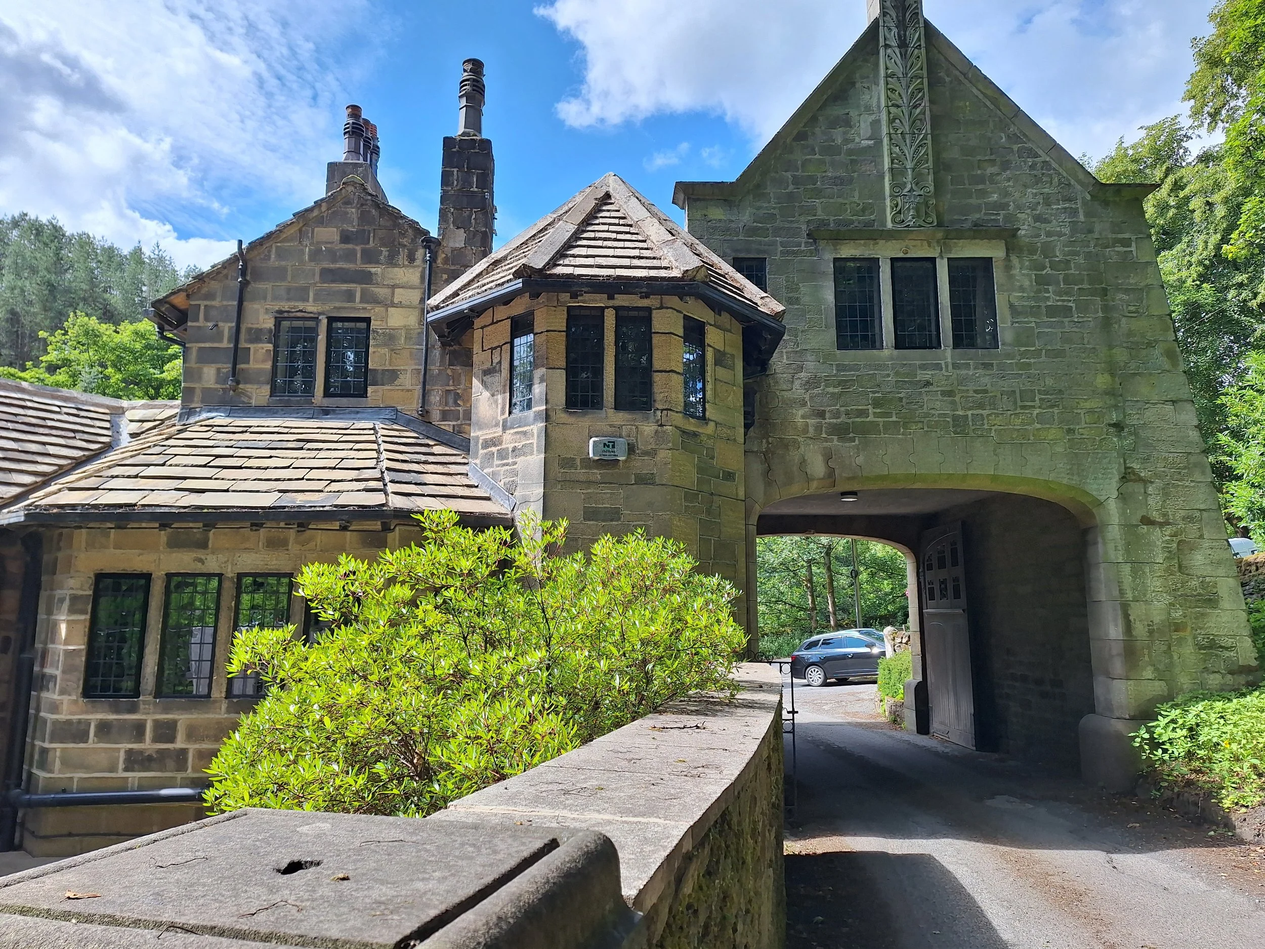 View of a stone house with a arched entrance, multiple windows, and a chimney, surrounded by green trees and shrubs on a sunny day.