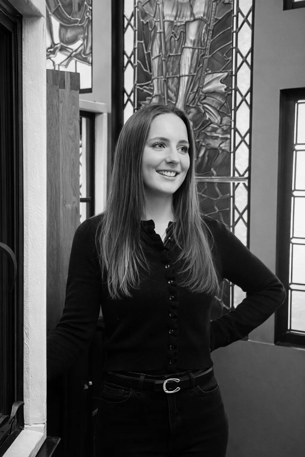 A smiling woman standing indoors near a door with a window, artwork with abstract designs in the background, wearing a black button-up shirt and black pants.