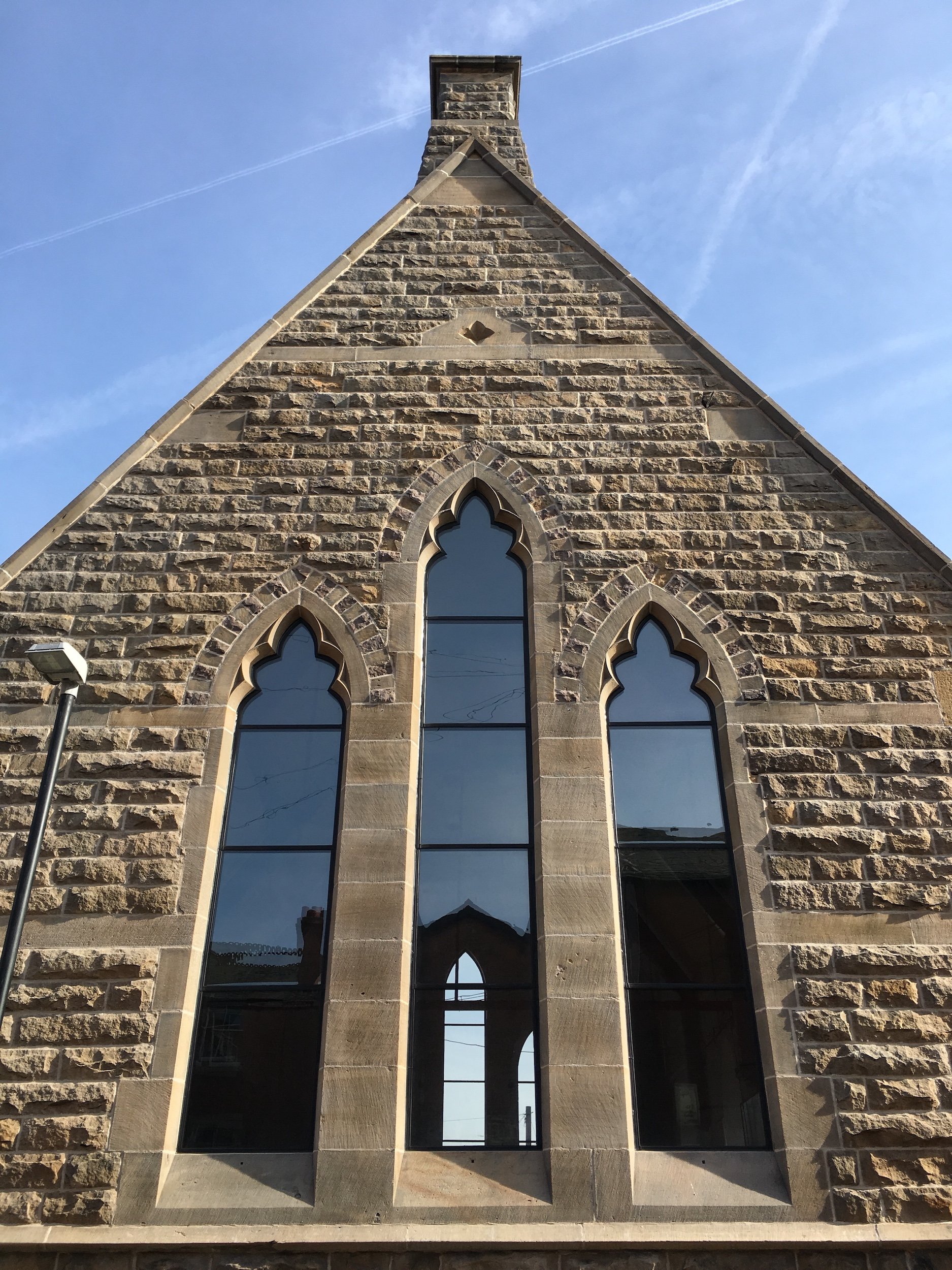 Front view of a stone church building with three tall gothic-style arched windows reflecting the sky, a chimney at the top, under a blue sky with a contrail.