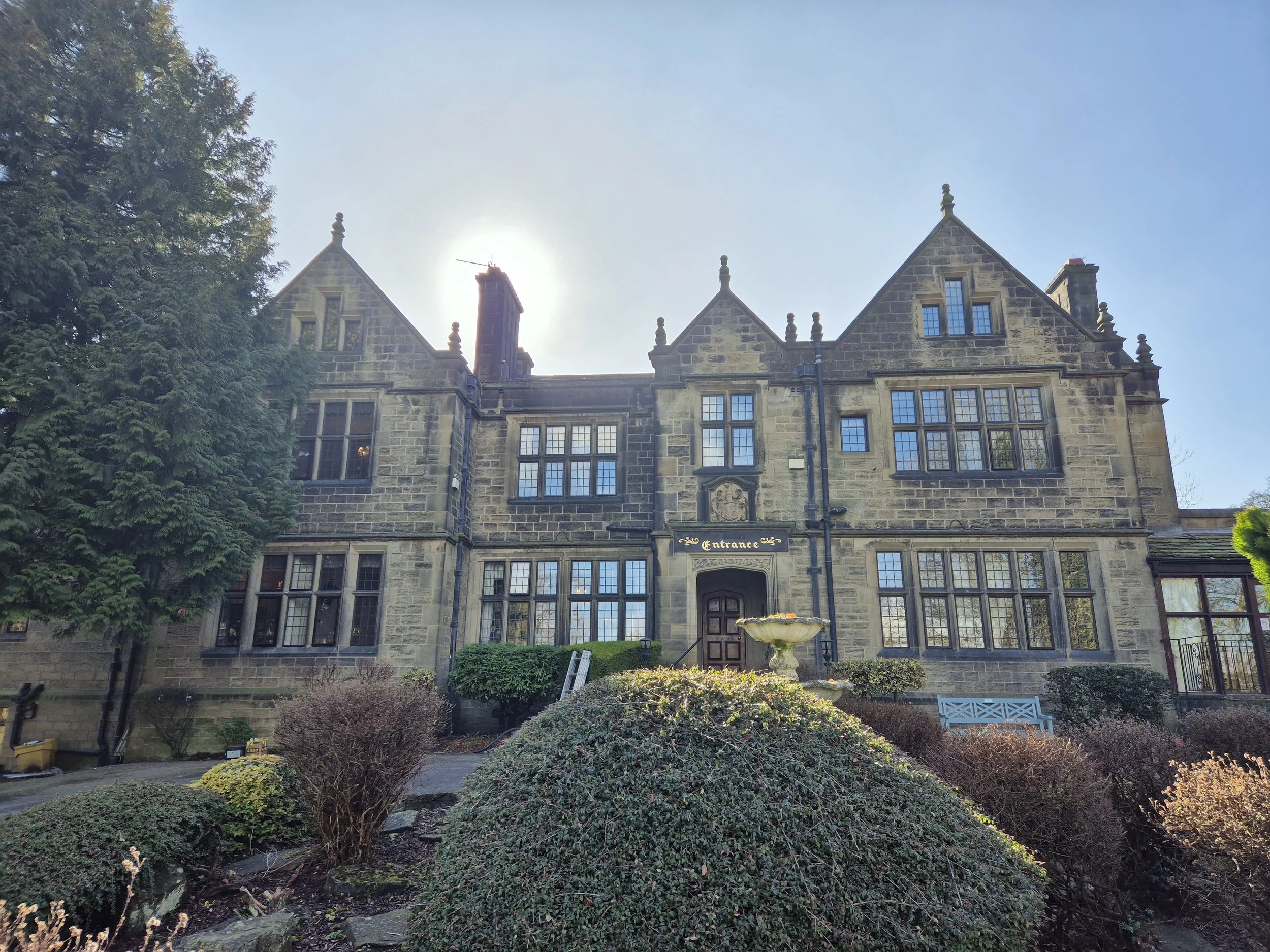 A large stone building with Gothic windows and a sign reading 'Entrance' above the door, surrounded by shrubs, trees, and a clear blue sky.