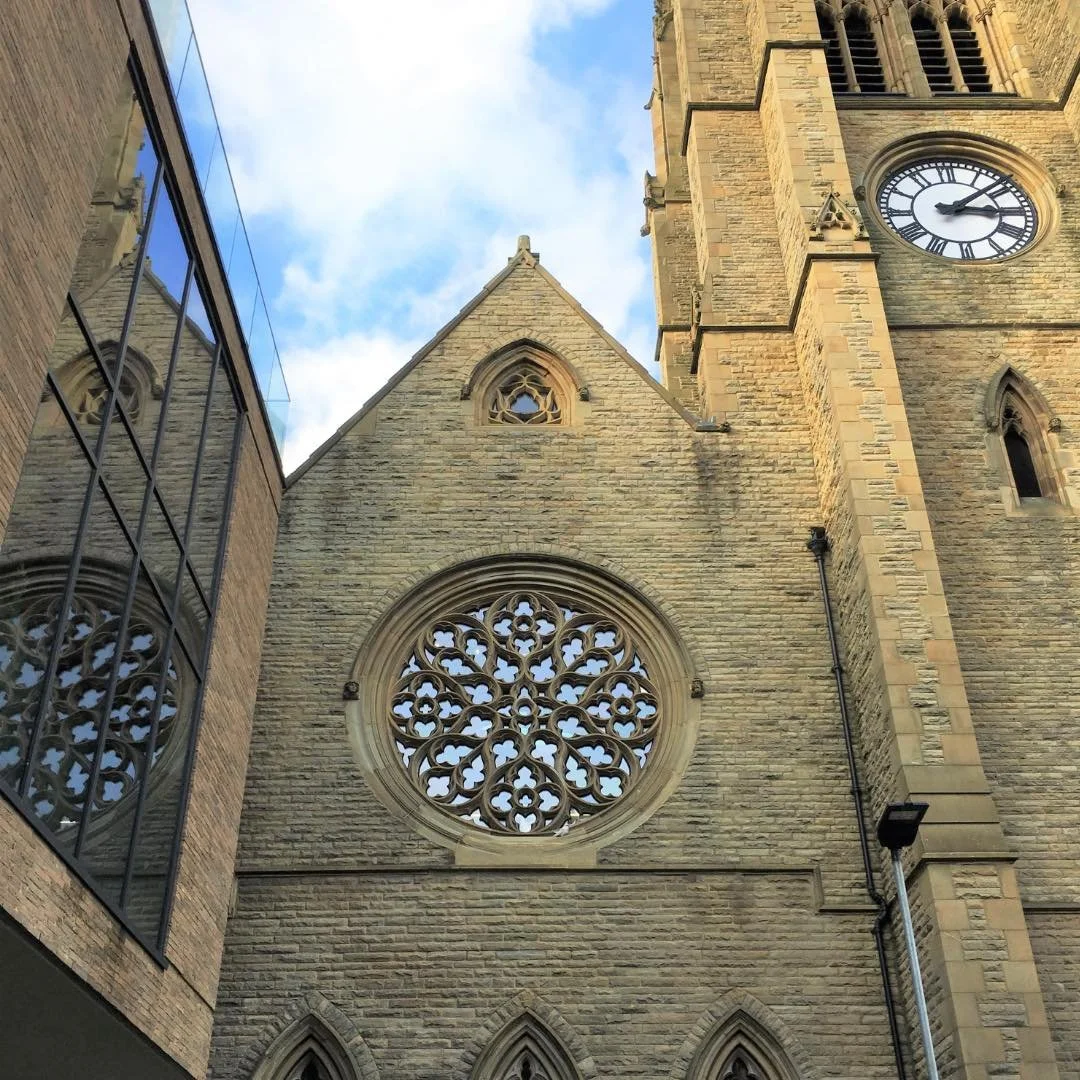 Close-up view of a historic stone church tower with a clock and intricate circular stained glass window, with a modern glass building reflecting in the side window.