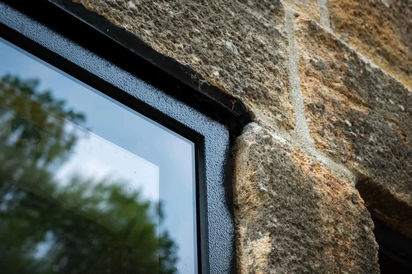 Close-up of a window corner with a black metal frame set into a weathered stone wall, with trees reflected in the glass.