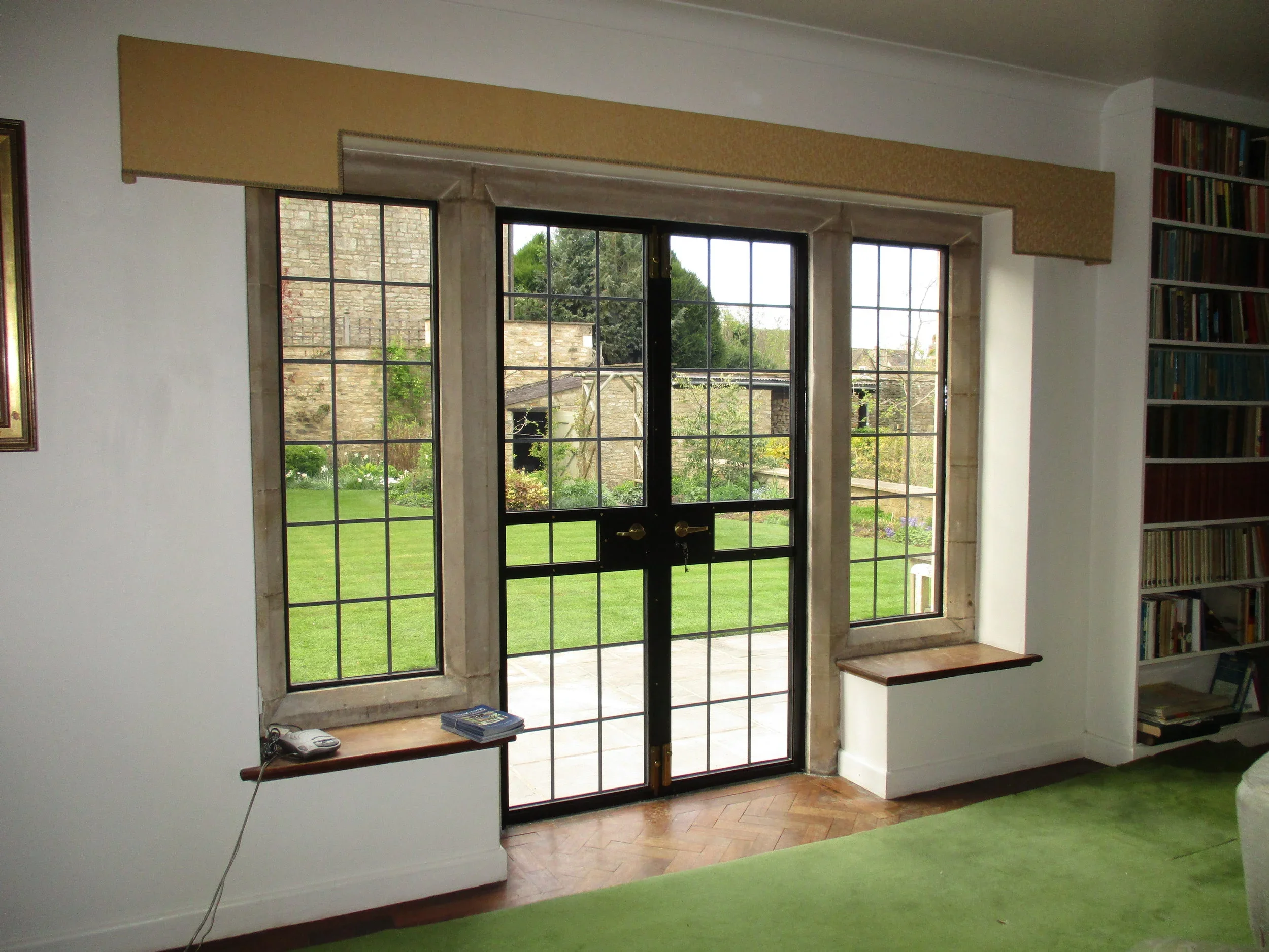 Interior view of a living room with large glass doors leading to a garden, flanked by two tall windows. There is a bookshelf on the right side and a green carpet on the floor.