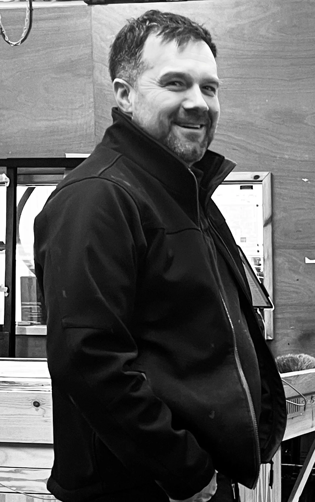 A man with short hair and a beard smiling, wearing a black jacket, in an indoor setting with wooden walls and posters or signs in the background.