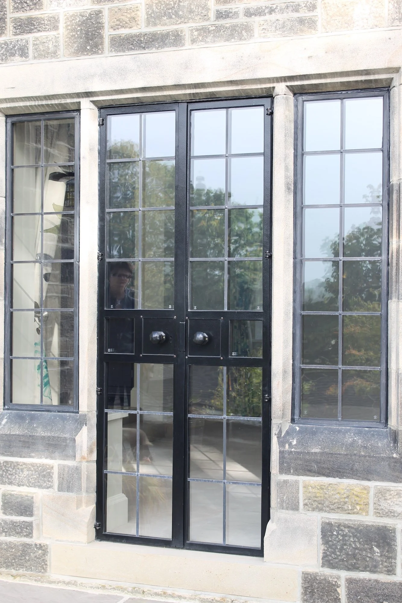 Black-framed double door with glass panels and decorative handles, flanked by two large glass windows with grid muntins, set into a stone building with brick and stone exterior.