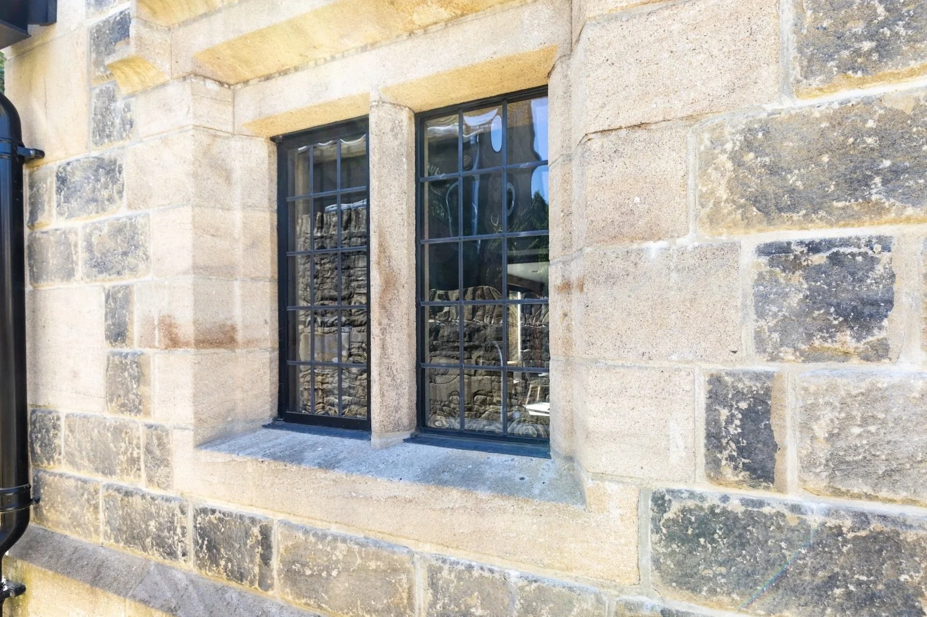 Close-up of a stone building wall with two rectangular windows with black wrought iron bars. The wall consists of large, uneven, weathered stones in shades of beige and gray. The windows have small panes and are set within the stone wall.