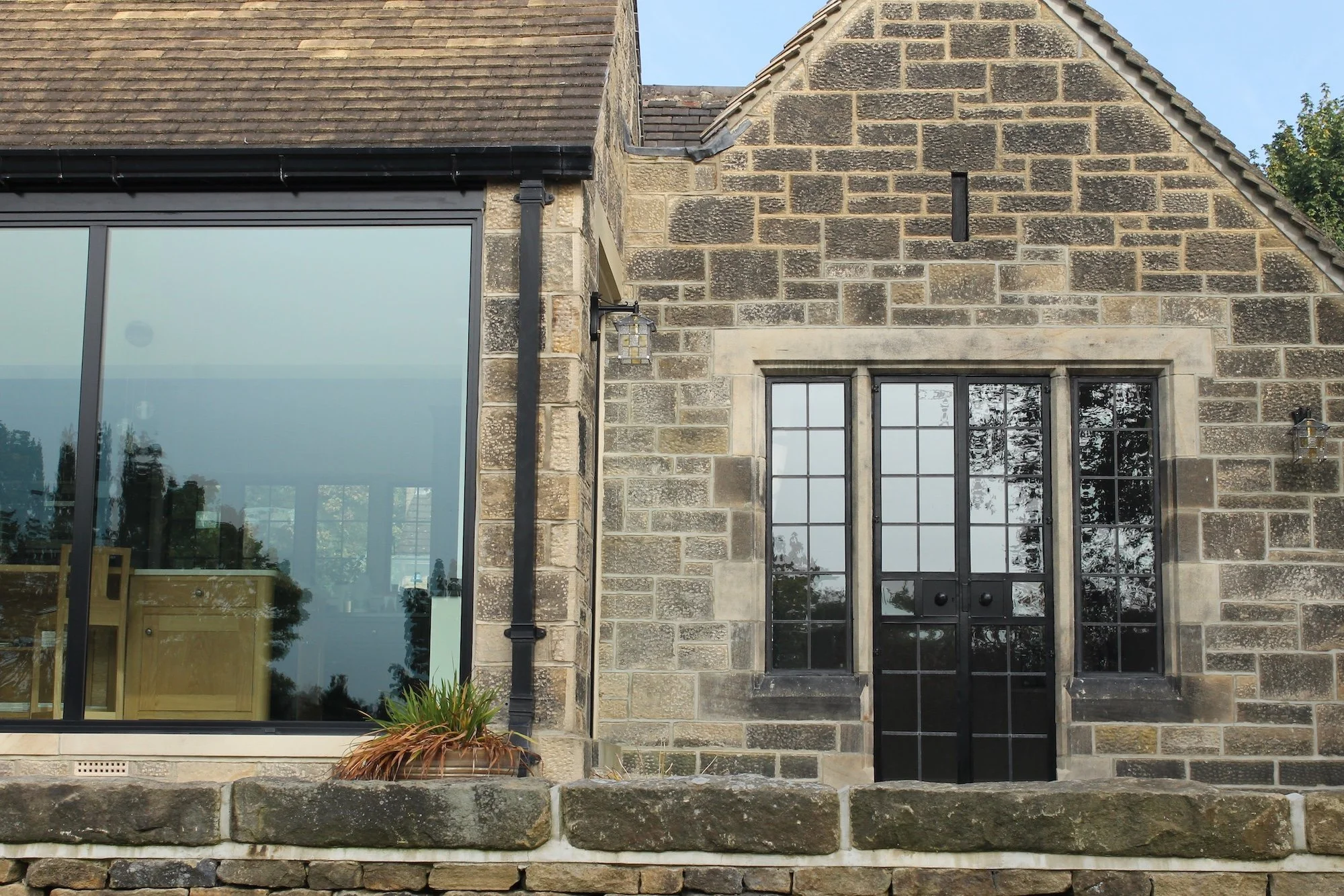 Close-up of a building's exterior showing a stone wall, black-framed windows, and a glass door, with a stone step and some potted plants in front.