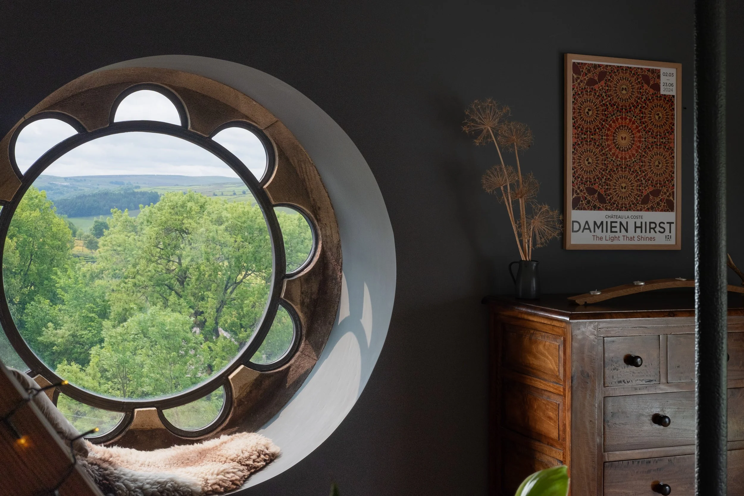 A round window with a flower-like shape cutout, showing a view of green trees and rolling hills outside, with a plush blanket on the windowsill. Inside, there is a dark wall, a wooden dresser, a black vase with dried wildflowers, and framed artwork b