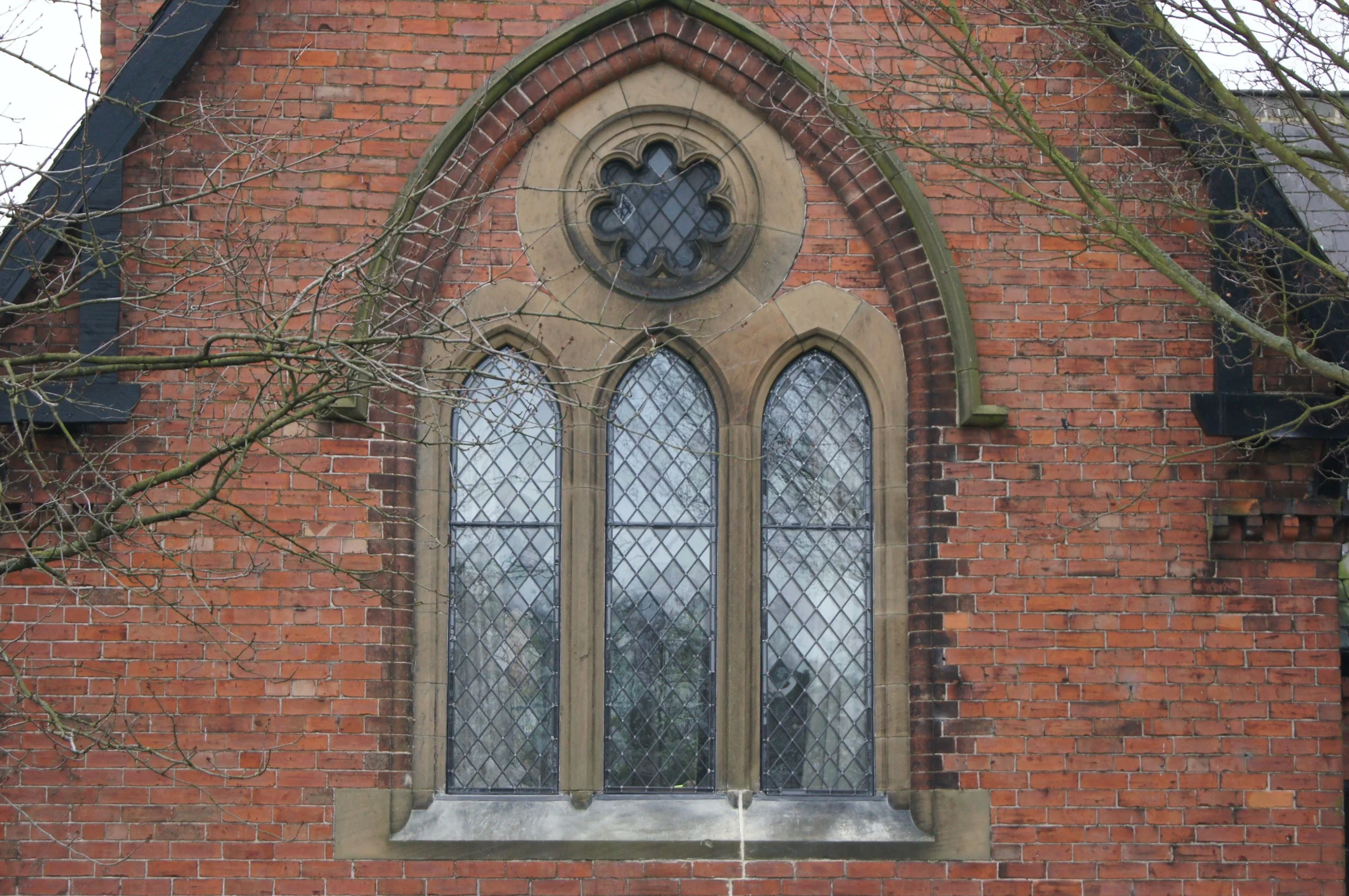 A brick church window with a pointed arch and decorative stone framing, surrounded by bare tree branches.