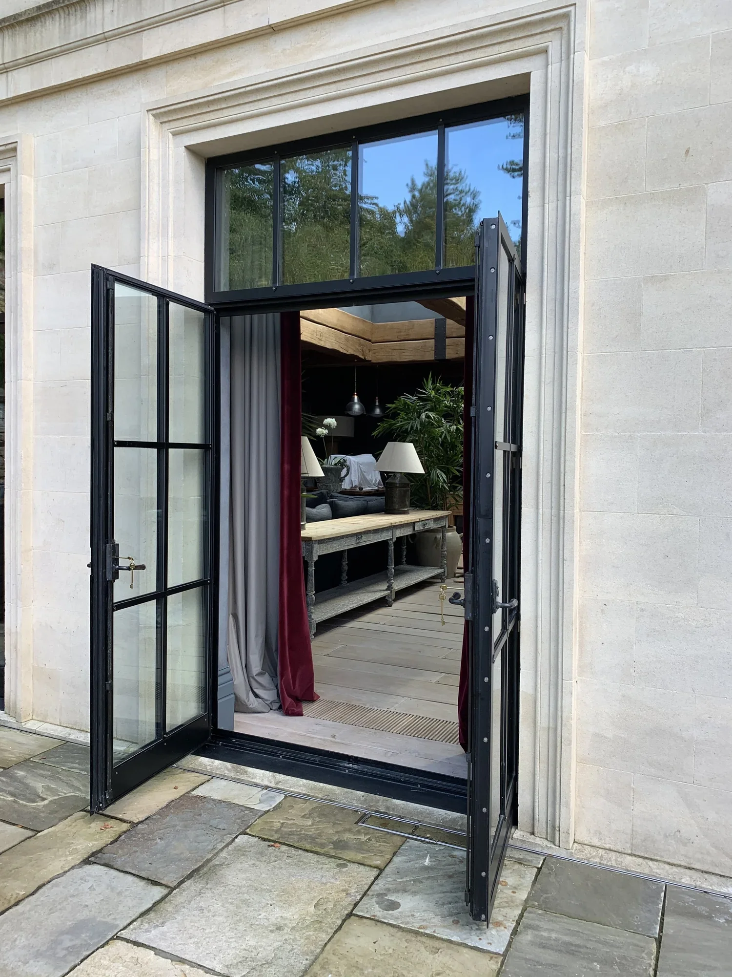 Open black metal patio door reveals a cozy indoor living space with a wooden floor, a large potted plant, a table with lamps, and a red curtain.