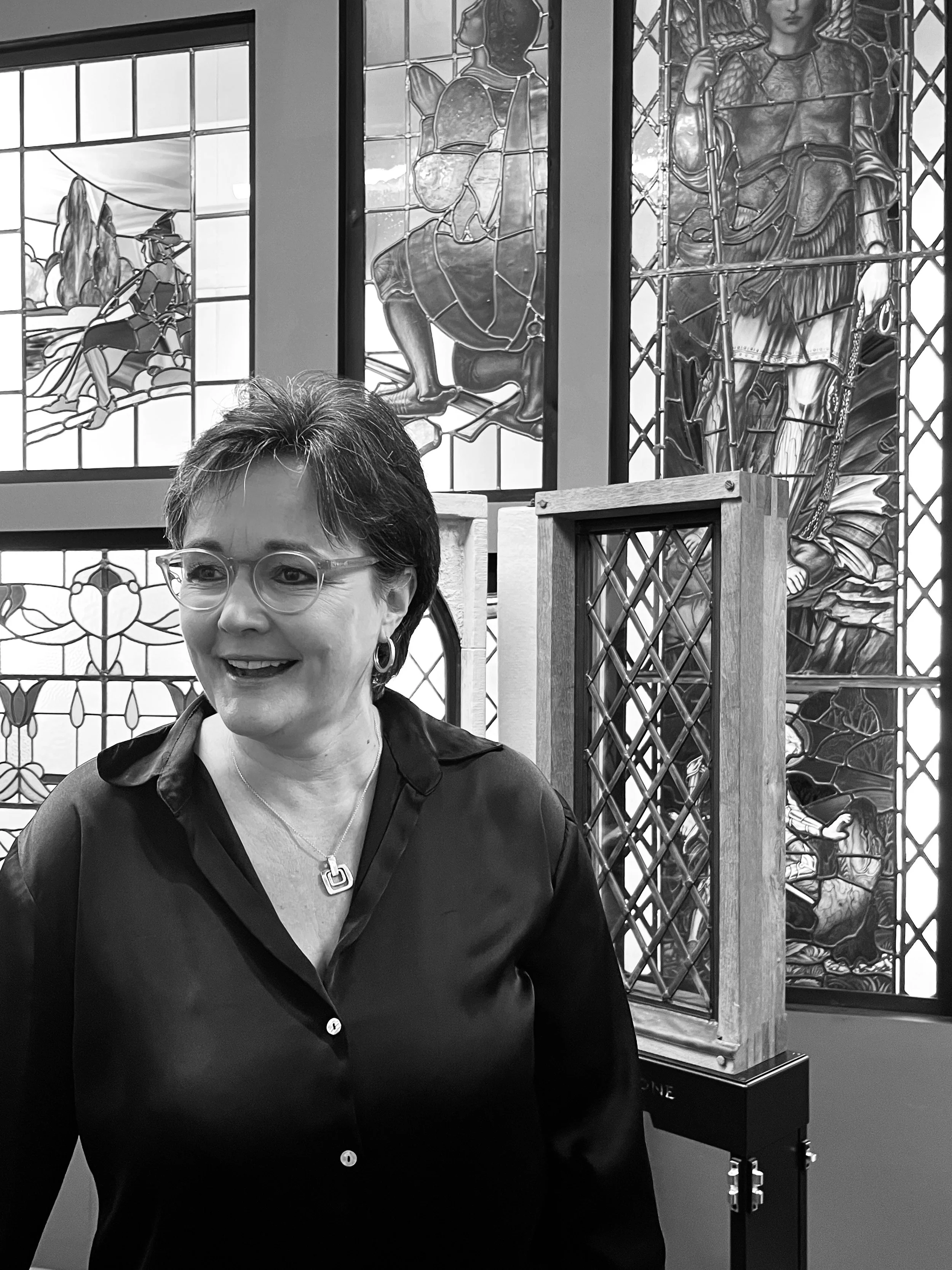 Black and white photo of a woman with short hair, glasses, earrings, and a necklace smiling in front of stained glass windows and an art installation of wooden frames with intricate designs.