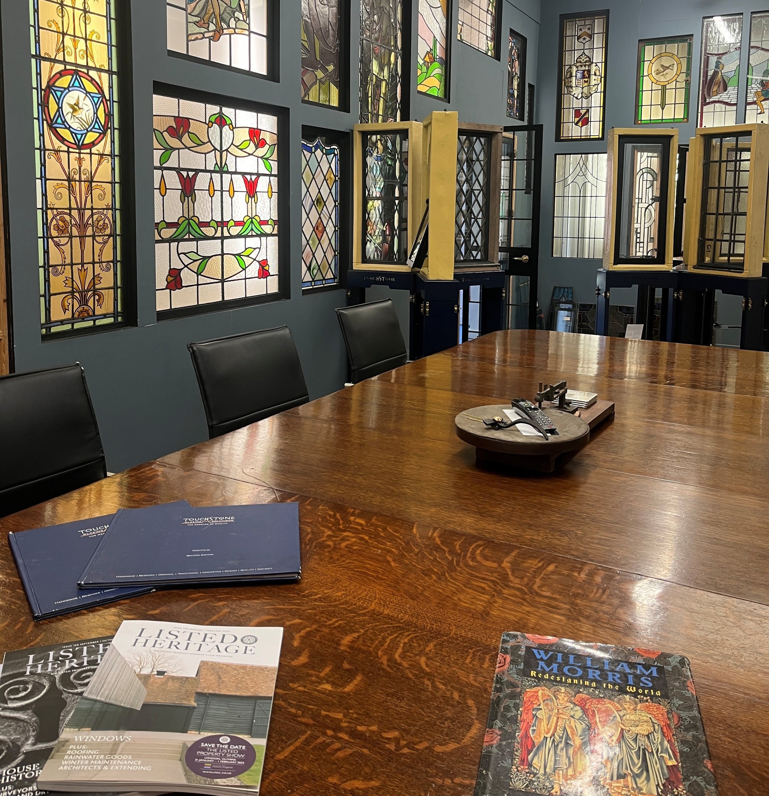 Display of books and magazines on a wooden table in a room decorated with stained glass windows and framed artwork on the wall.