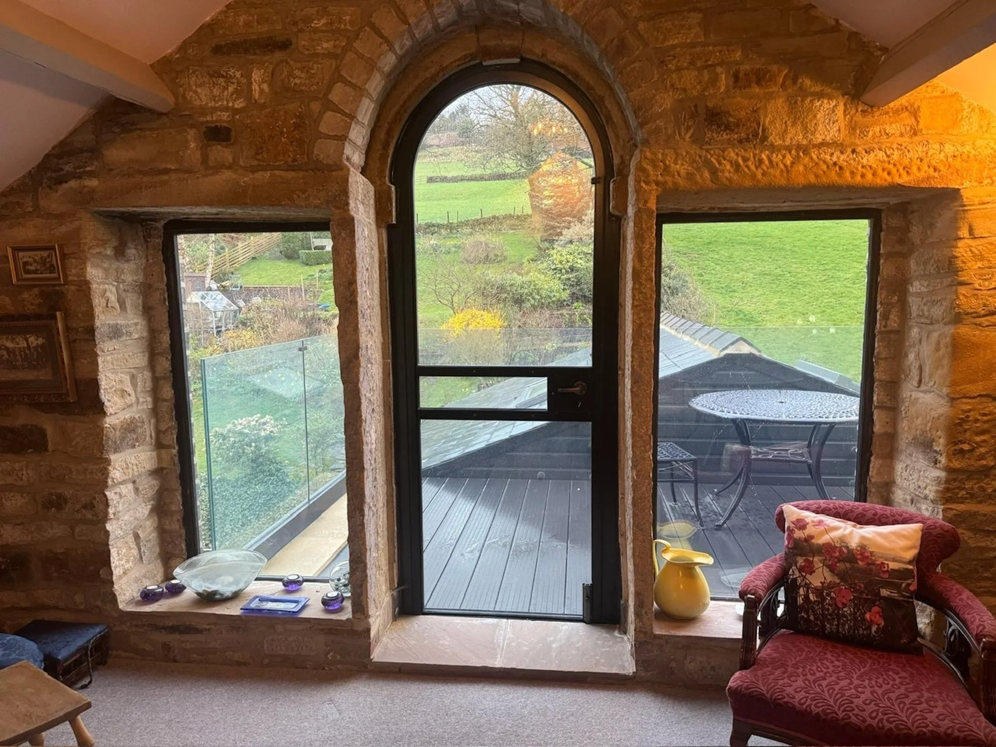 View of a living space with a stone wall and large arched window overlooking a green outdoor space. The room features a small patio with black chairs outside, and interior decor including a red patterned armchair, a yellow vase, and decorative glass 
