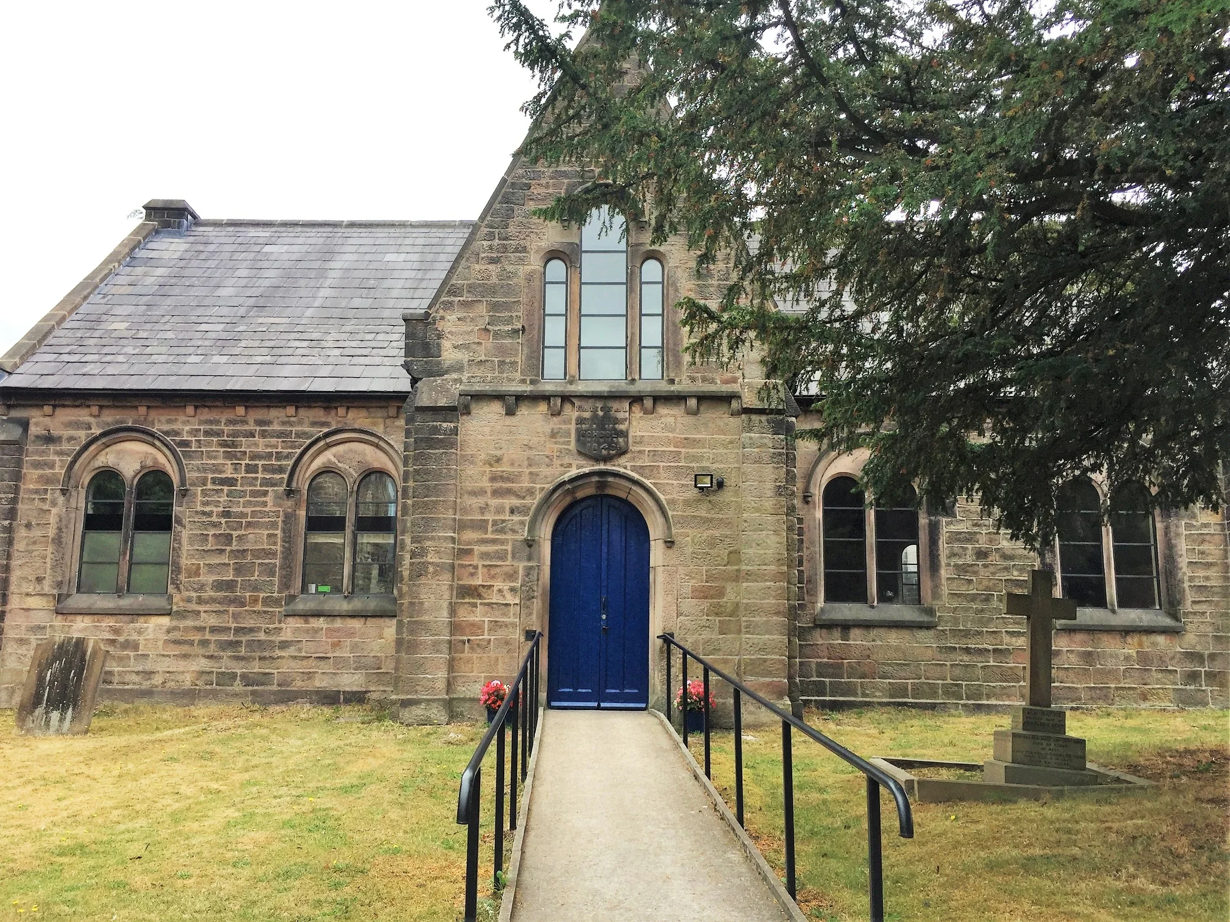 A stone church with a pointed roof and tall, arched windows. A blue door is accessible via a small ramp with a handrail. There are potted flowers near the doorway, and a large tree partially obscures the right side of the church. The sky appears over