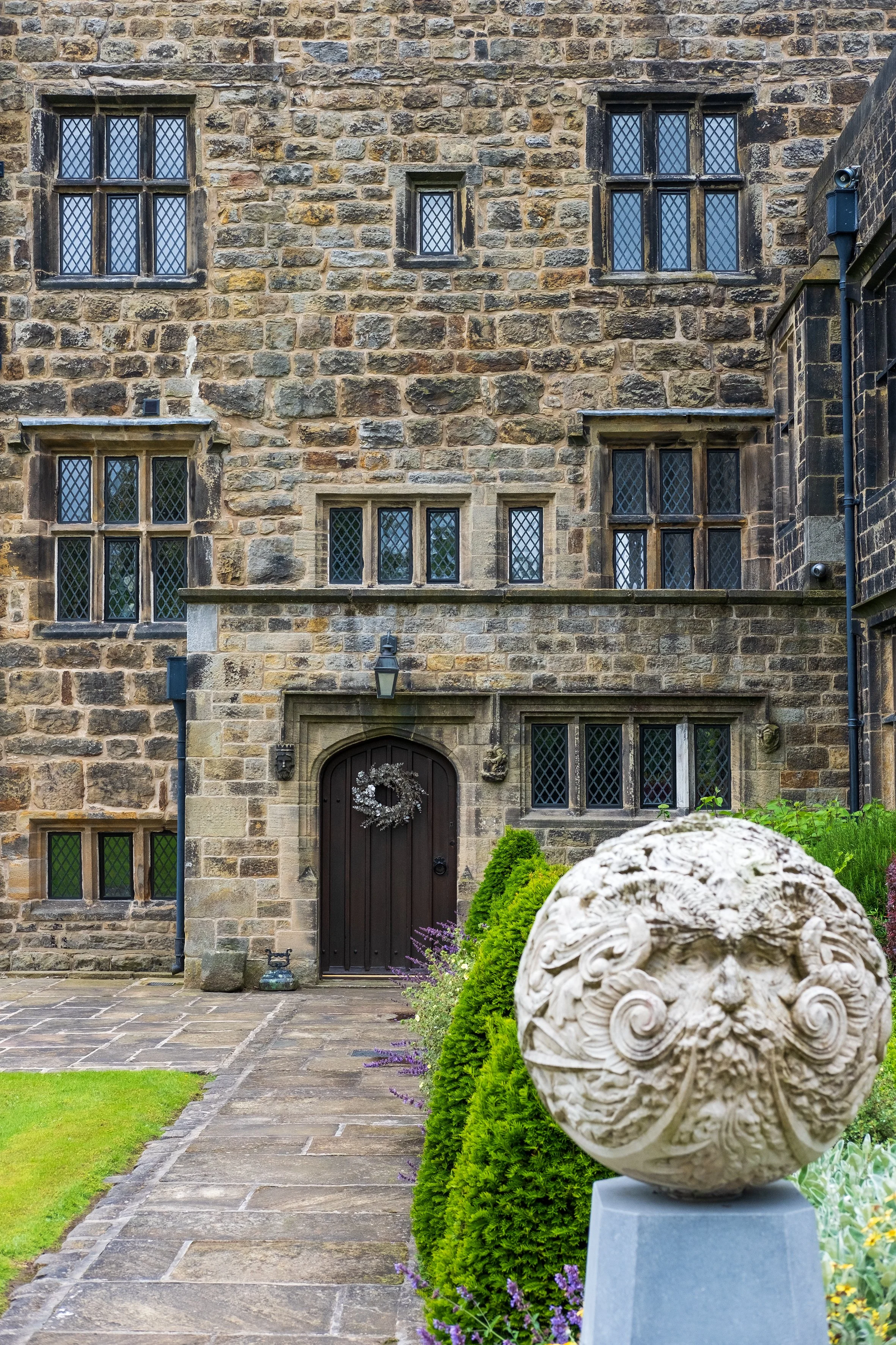 Stone building with multiple small and large windows with diamond-shaped panes, wooden door with wreath, stone pathway, and decorative stone sculpture in a garden with greenery and purple flowers.