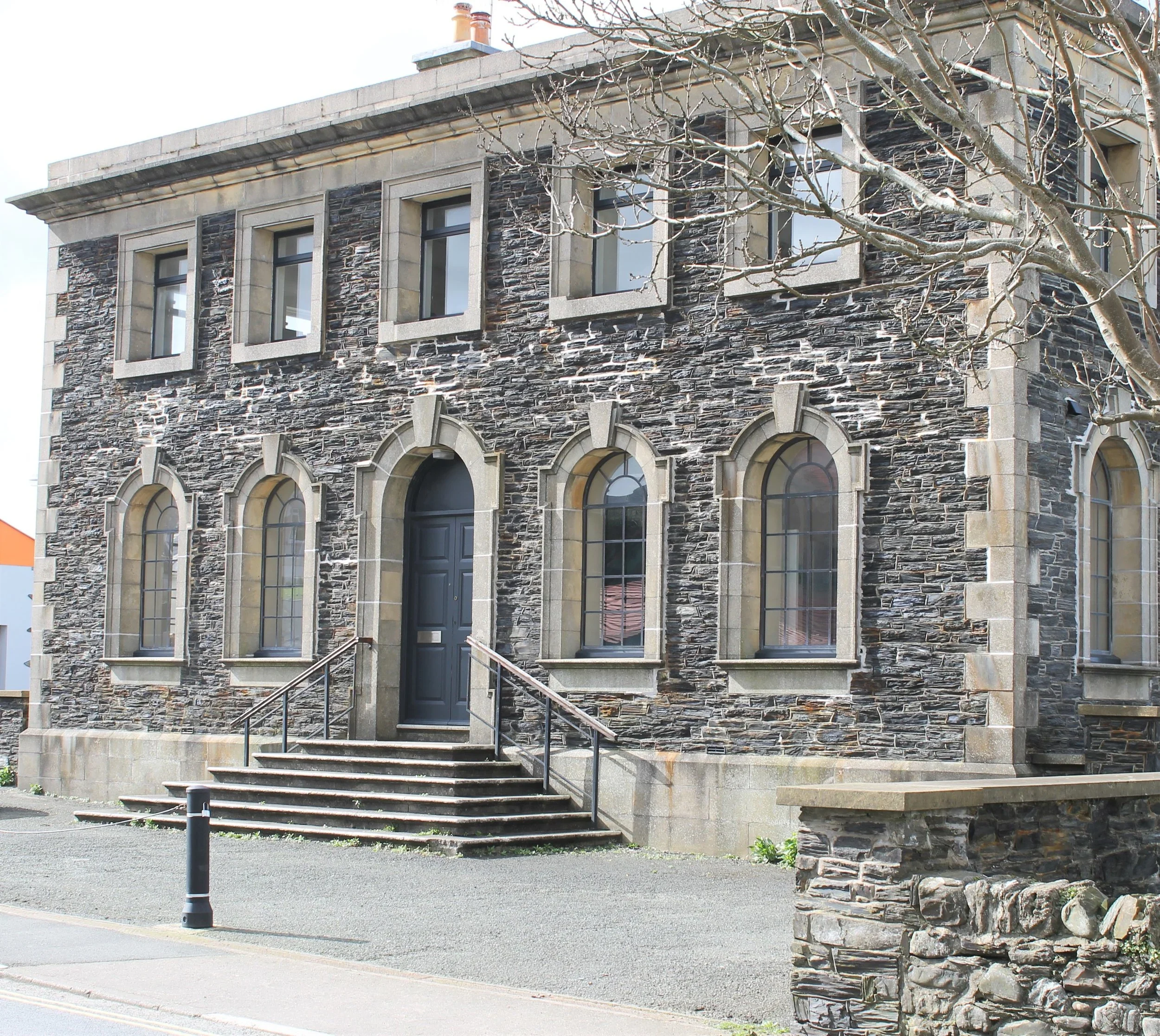 A historic stone building with arched windows and a central door, accessible via stairs, situated on a city street.