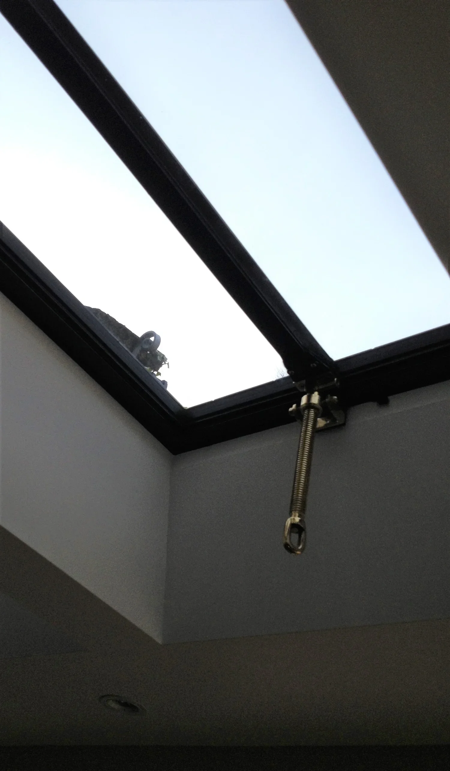 Interior view of a ceiling with an open skylight window and a metal spring loaded latch hanging from the window frame.