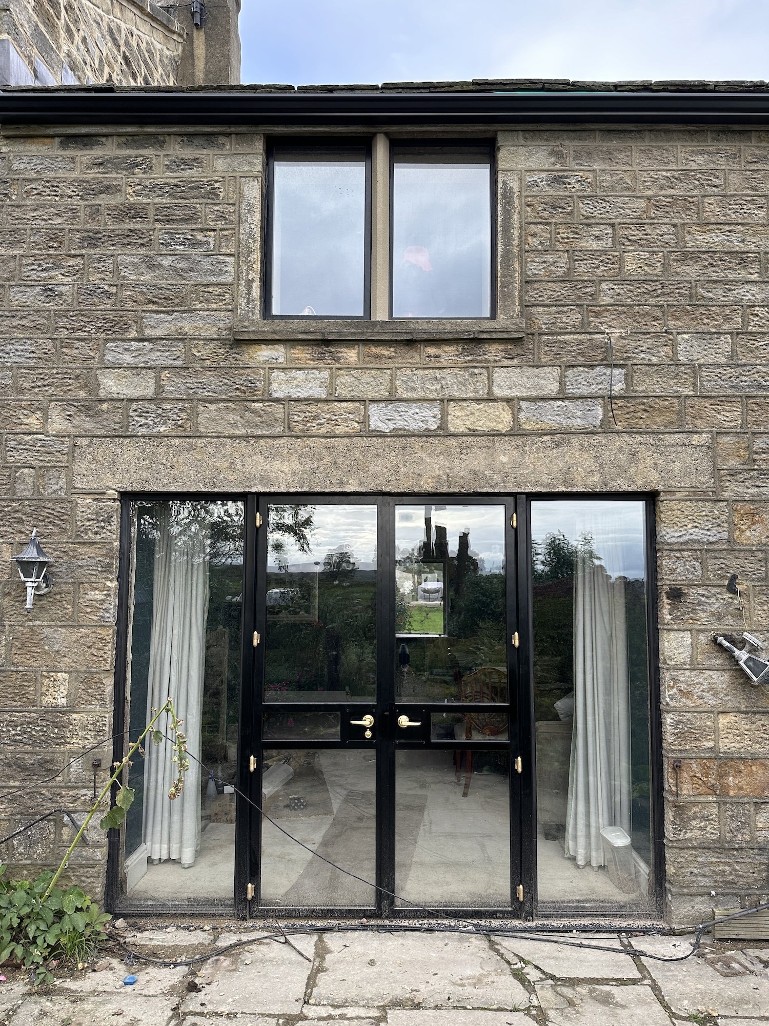 The image shows a set of black-framed sliding glass doors leading into a house, with brick walls on either side and a stone lintel above. The interior is partially visible, revealing a chair and some furniture. Outside the doors, there are some plant