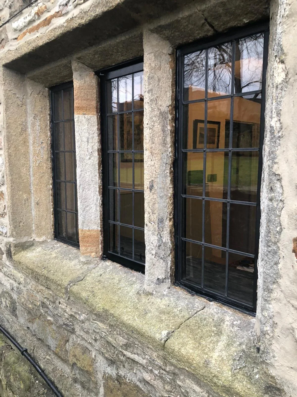 Three window panes with black frames and divided glass, embedded in a rustic stone wall with weathered stone sill, overlooking an interior with framed artwork and outdoor trees reflected in the glass.