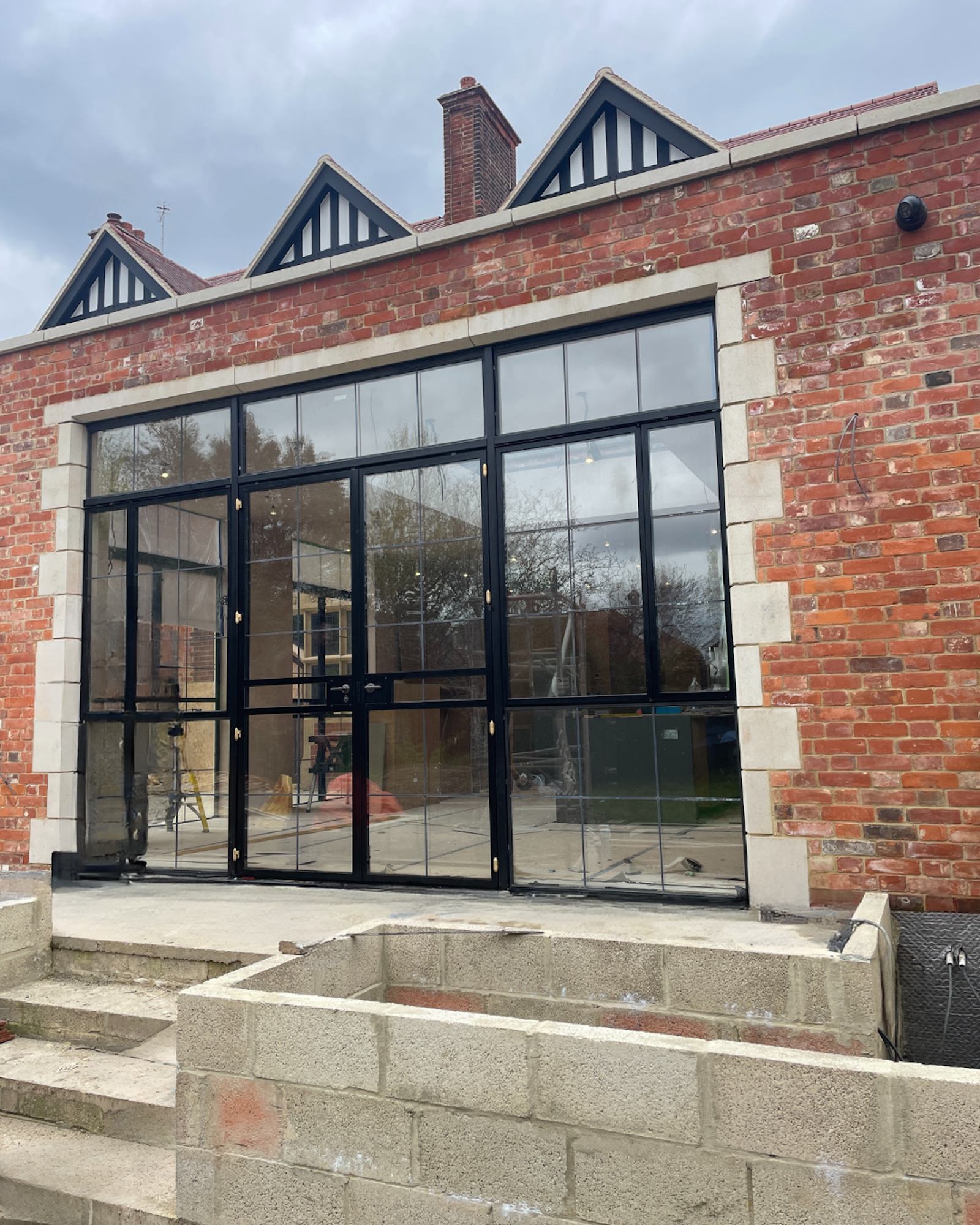 Building under construction with brick walls, large black-framed glass doors, and a concrete staircase and foundation in the foreground.