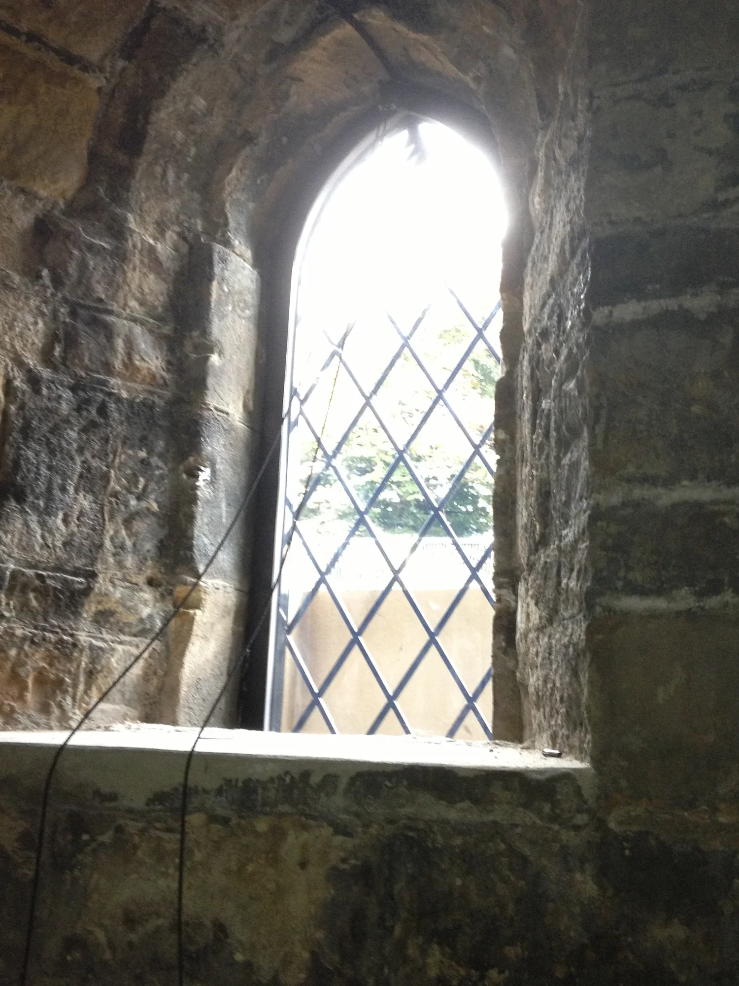 View through a small stone window with a metal grille, showing bright outdoor scenery with trees.