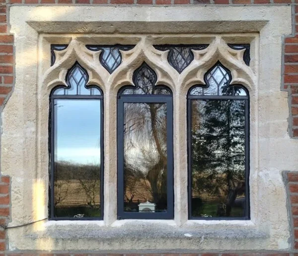A window with Gothic-style stone tracery in a brick building, with reflections of trees and a cloudy sky in the glass.