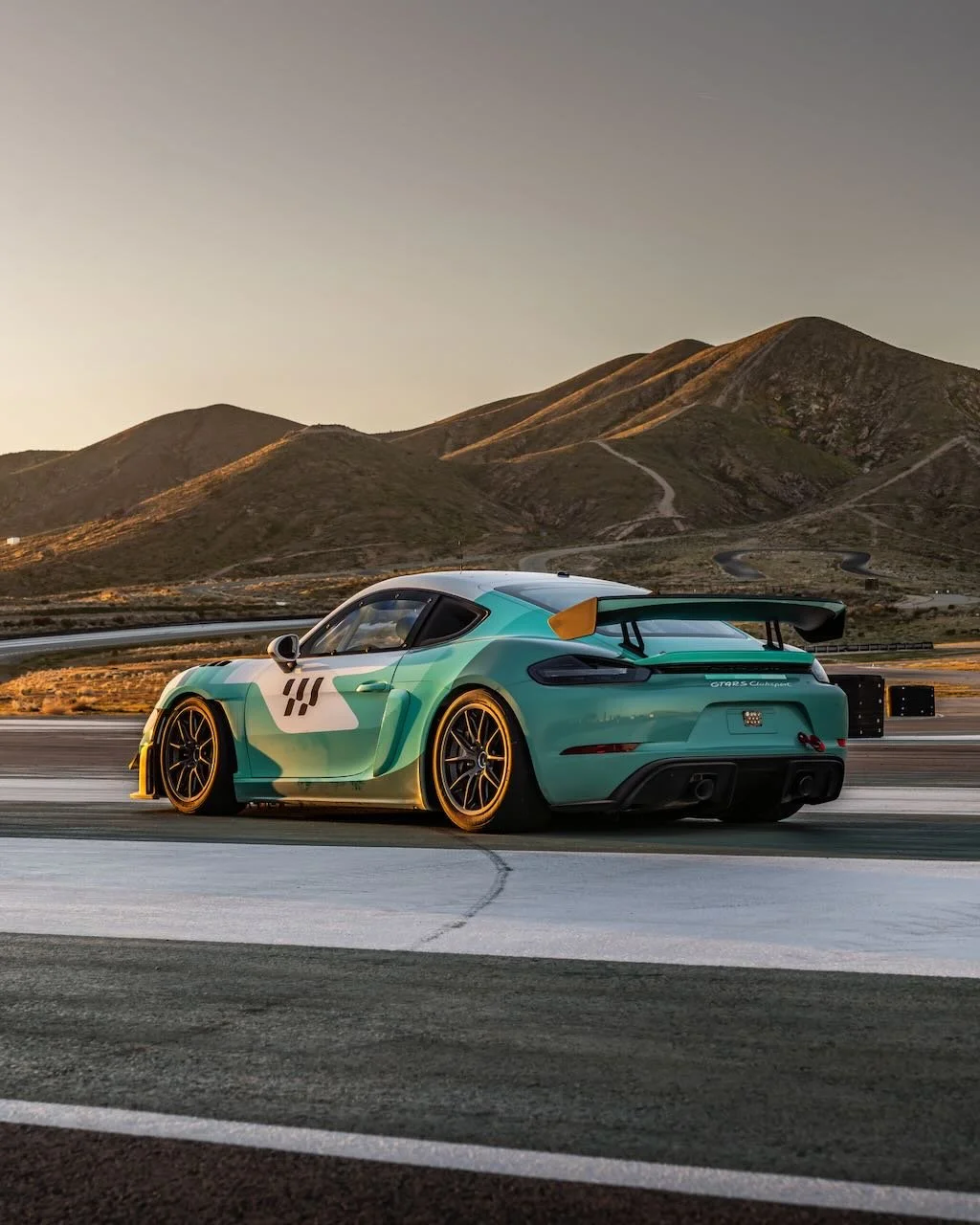 A turquoise Porsche 911 GT2 RS Clubsport race car driving on a racetrack in a mountainous area during sunset.