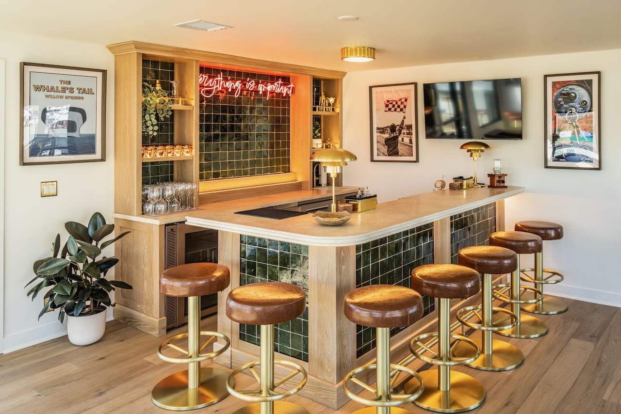 Modern racetrack bar with a curved wooden counter, six leather and gold barstools, black tile accents, framed artwork on the walls, and a neon sign.