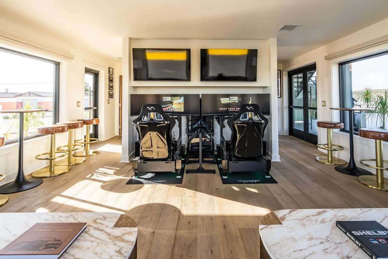 Room with two racing simulator setups, four flat-screen TVs mounted on the wall, and windows with outdoor views of racetrack.