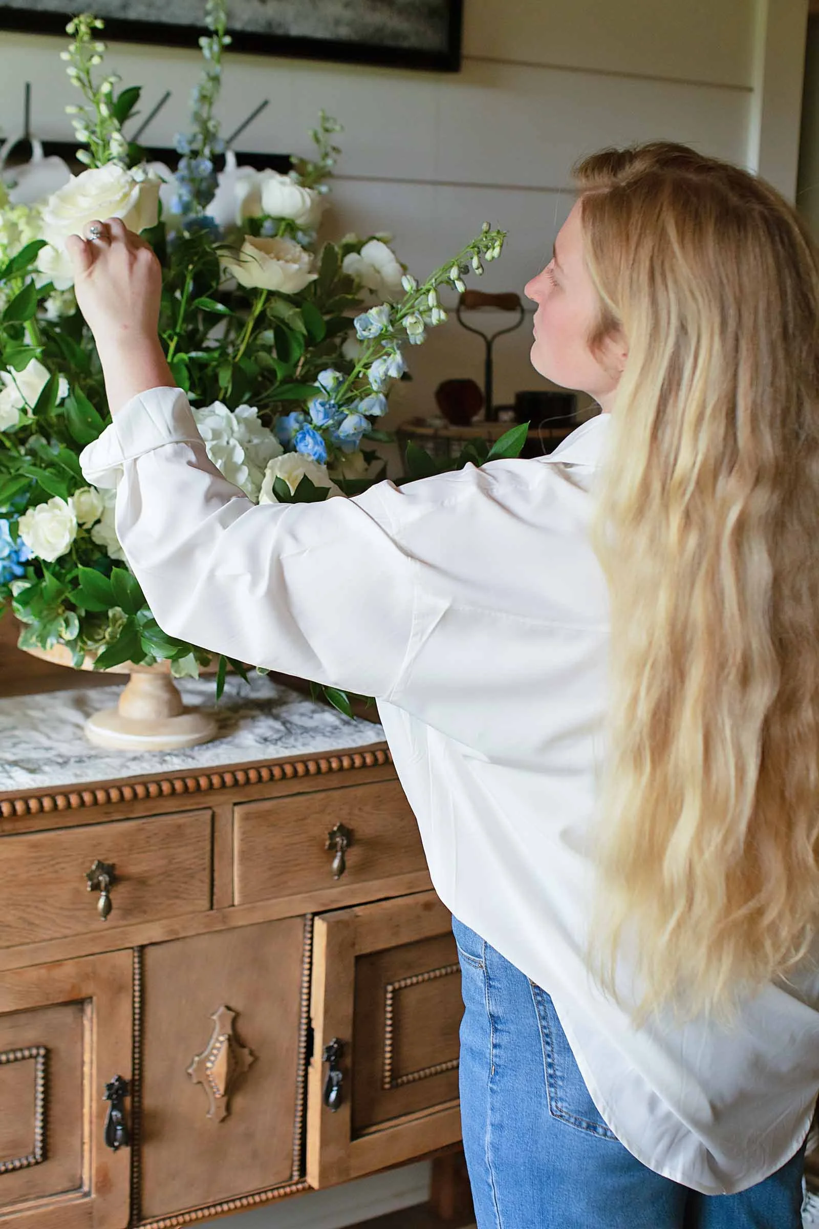 A  wedding florist in a white shirt arranging white and blue flowers in her studio in central Louisiana.