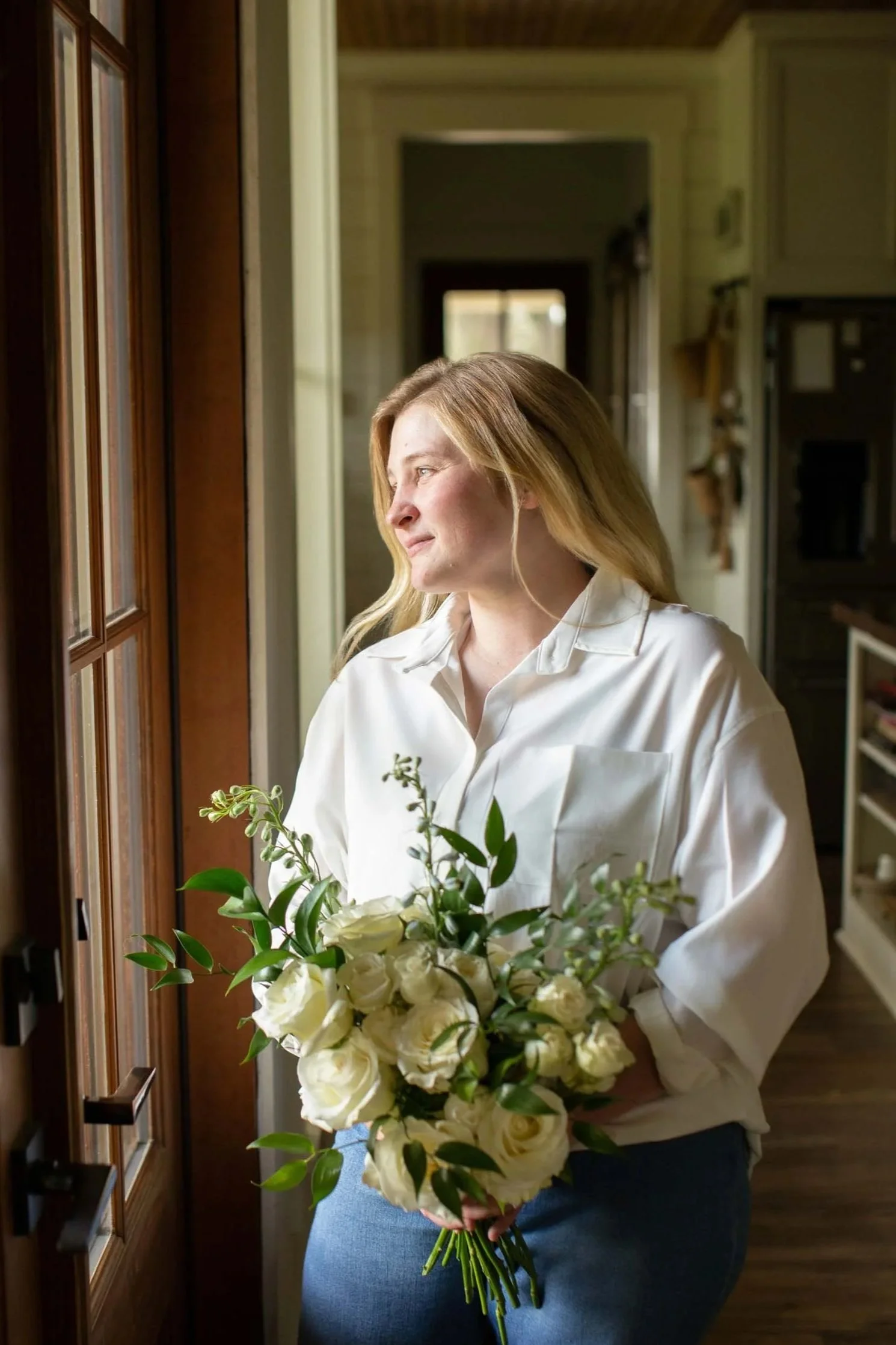 A central Louisiana florist holding a wedding bouquet, gazing out of a wood-framed window.