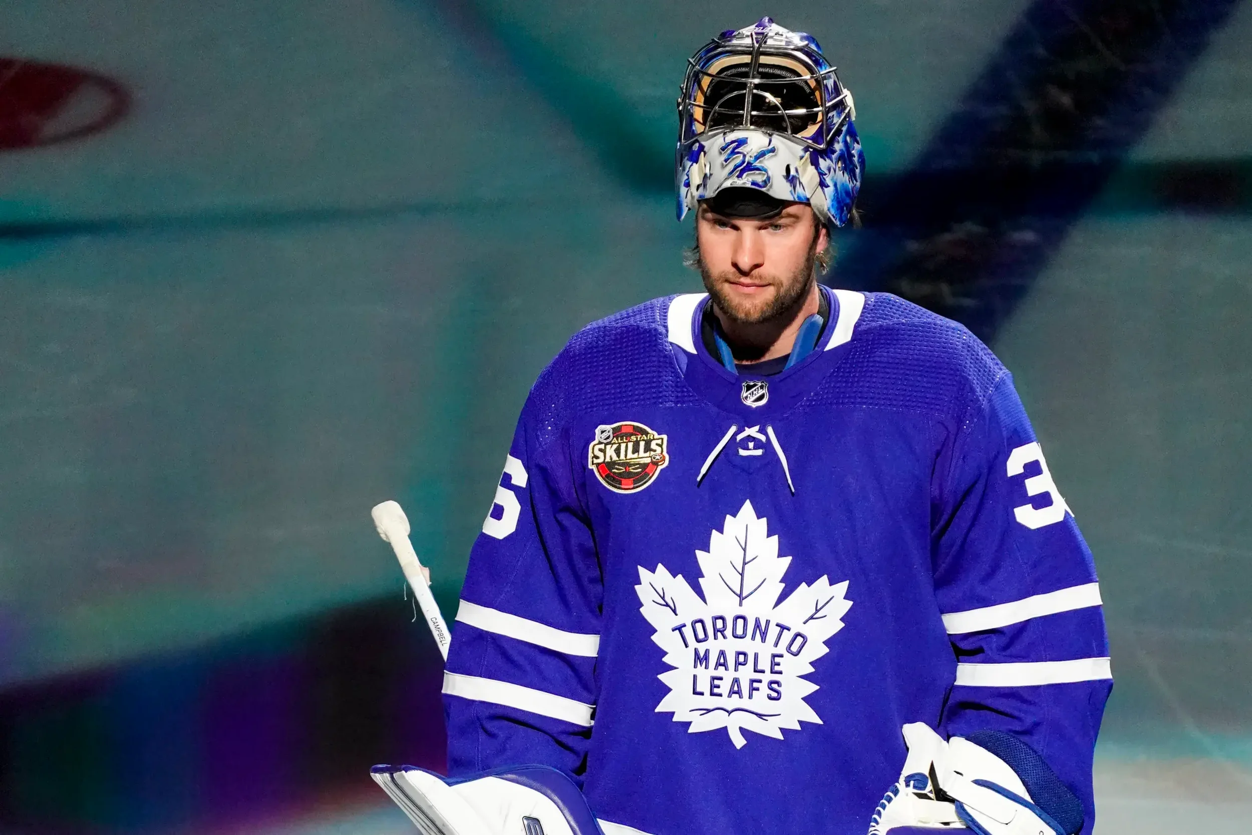 A hockey player wearing a blue Toronto Maple Leafs jersey with a white maple leaf and the team's name on it, a helmet, and goalie pads, standing on the ice during a game.