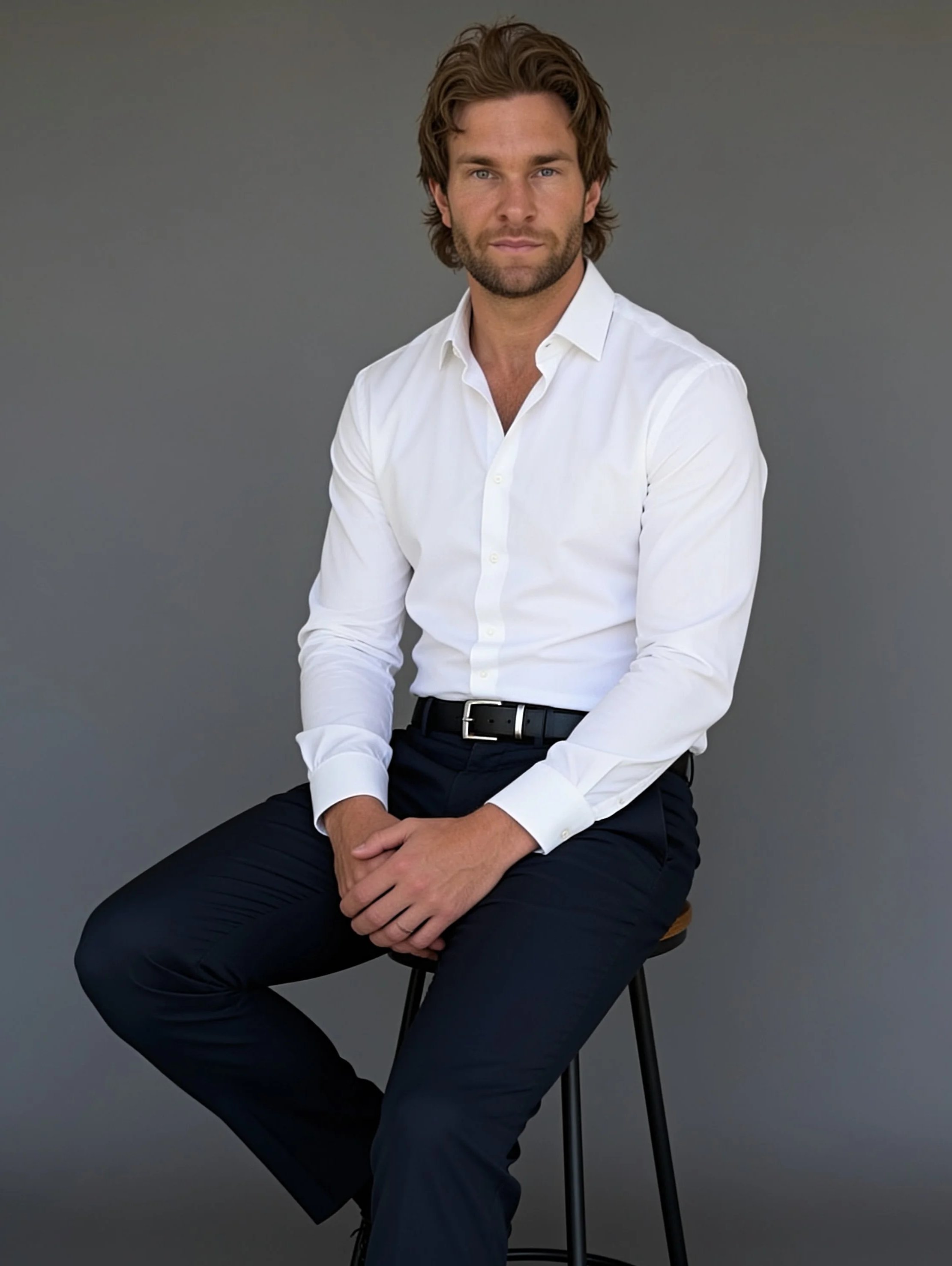 A man with brown hair and a beard, wearing a white dress shirt and dark pants, is sitting on a stool against a plain gray background.