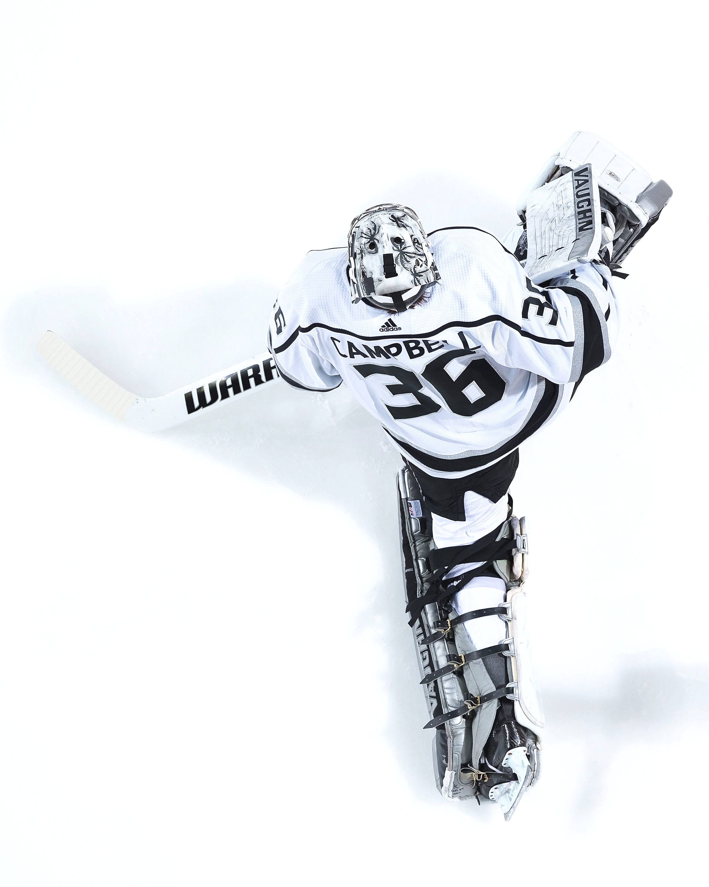 Overhead view of a hockey goalie in a white jersey with black details, kneeling on the ice with equipment, looking up. The jersey has the name 'CAMPBELL' and the number 30. The goalie gear includes a glove, blocker, leg pads, and a helmet, with a white glove and black leg pads, against a white ice surface.