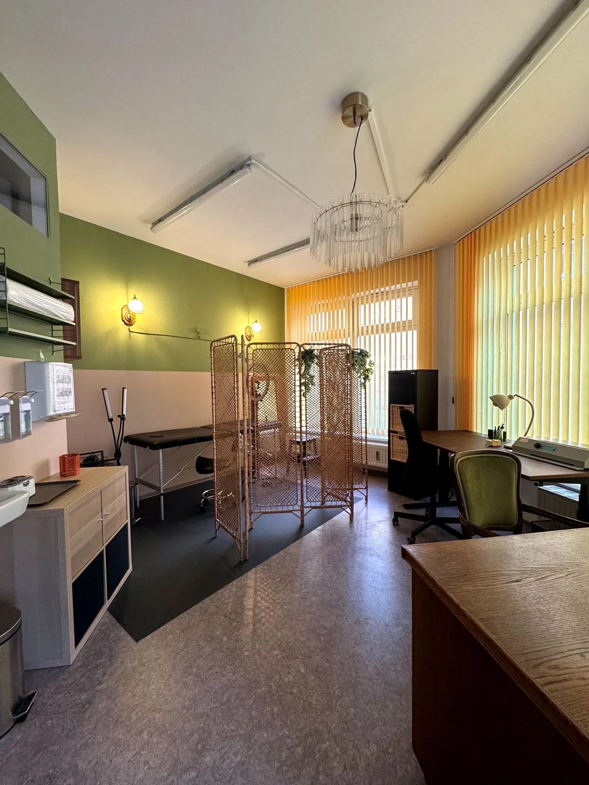Ein Raum mit grüner Wand, vertikalen gelben Jalousien an den Fenstern, einem wiederverwendbaren Raumteiler aus Rattan, einem Schreibtisch mit einem Computer und einem Schreibtischlampe, einem medizinischen Untersuchungstisch, medizinischen Geräten, und Beleuchtungskörpern an der Decke.