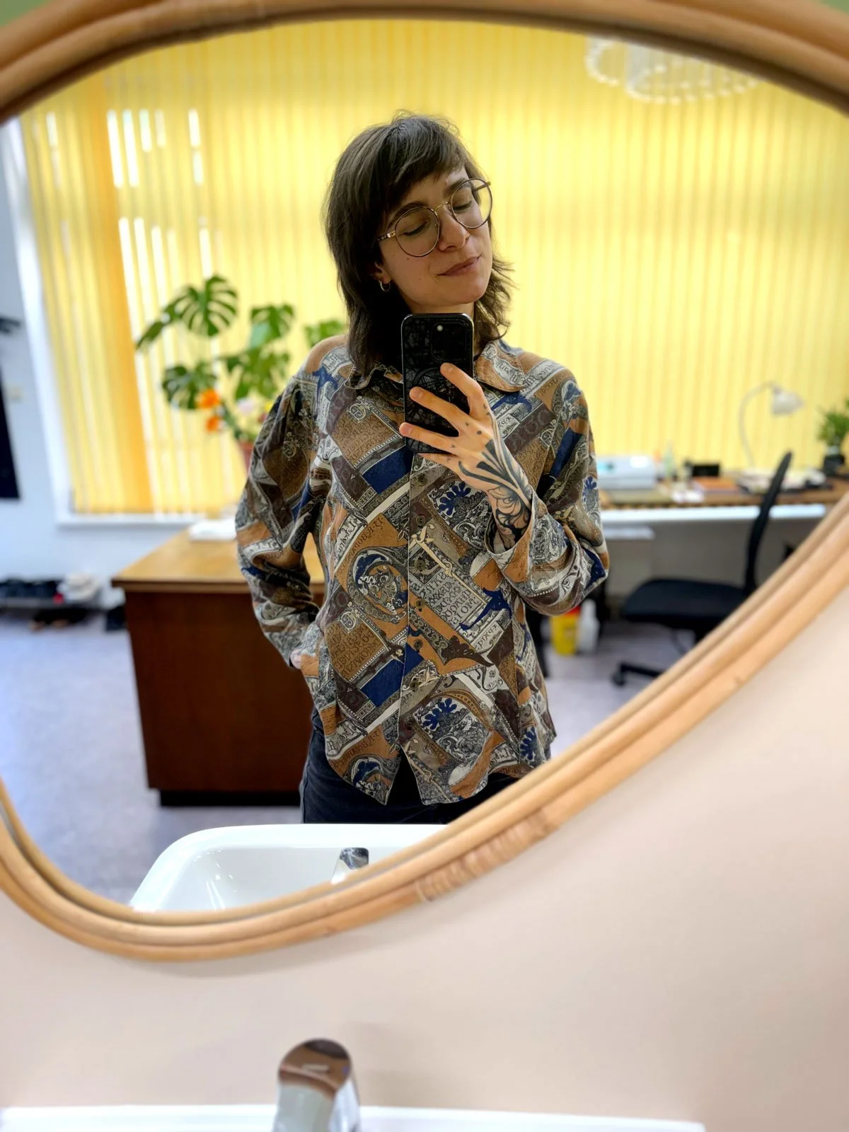 Frau macht Selfie in einem Büro mit gelben vertikalen Vorhängen, einer Pflanze und einem Schreibtisch im Hintergrund, und betrachtet sich in einem runden Spiegel.