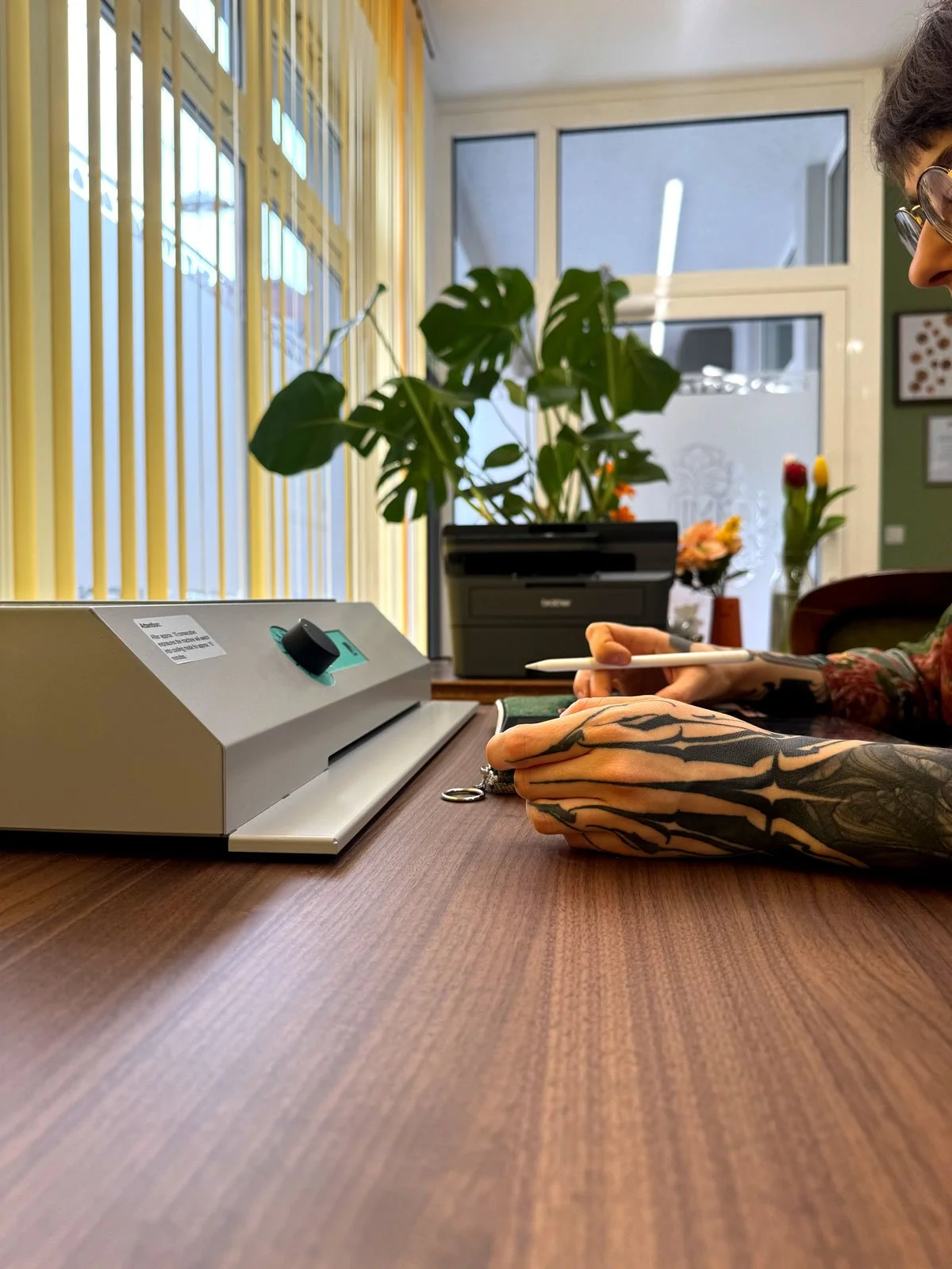 Ein Tisch mit einem Drucker, einer Hand mit Tätowierungen hält einen Stift. Im Hintergrund sind Pflanzen und große Fenster.