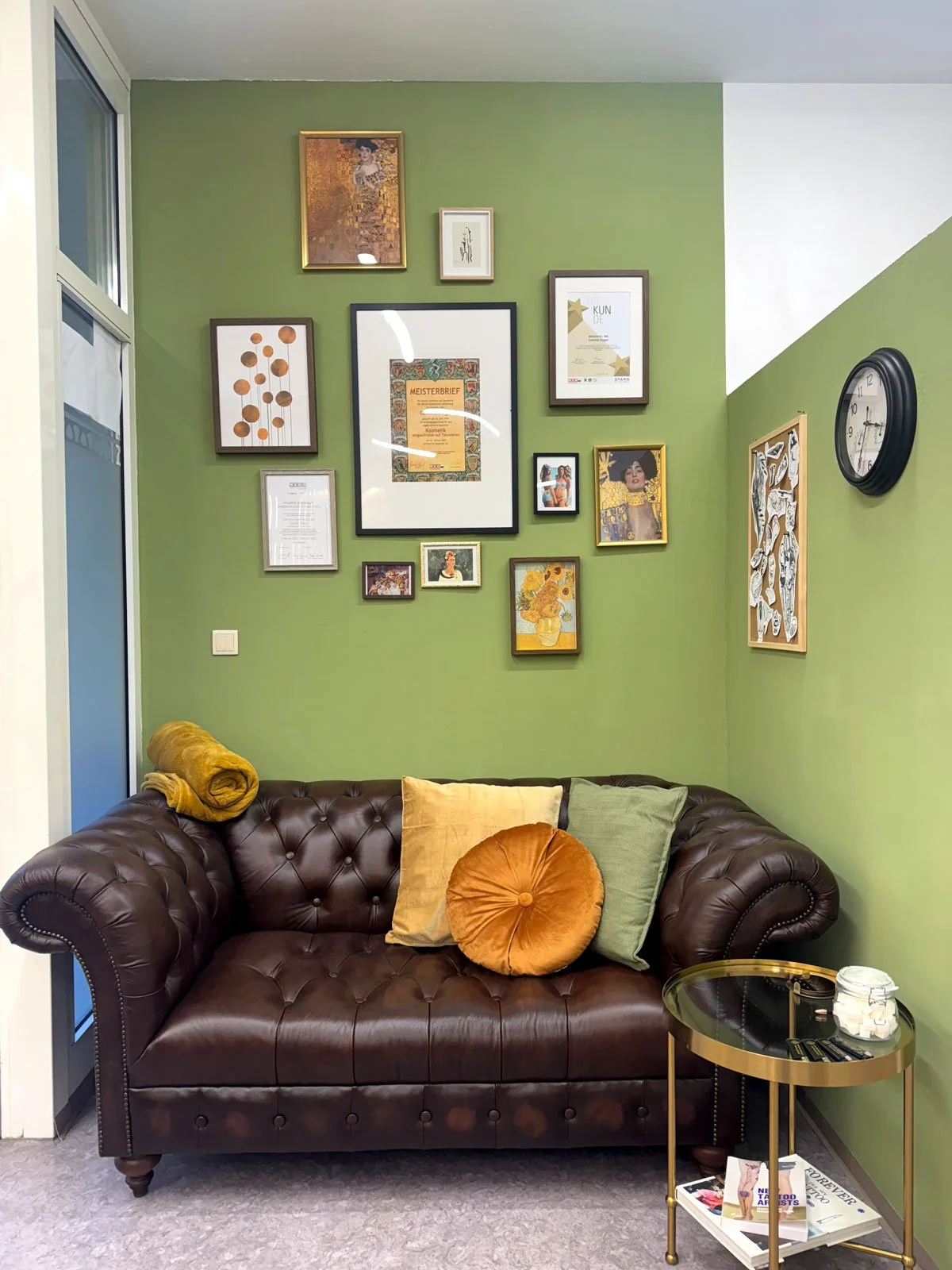 Gemütliches Wohnzimmer mit dunklem Ledersofa, bunten Kissen und einer grünen Wand mit mehreren Bilderrahmen und Kunstwerken. Es gibt eine kleine goldene Beistelltisch, auf dem eine Schüssel mit Zuckerwürfeln, eine Fernbedienung und eine Zeitschrift liegen. Ein Wandclock ist sichtbar.