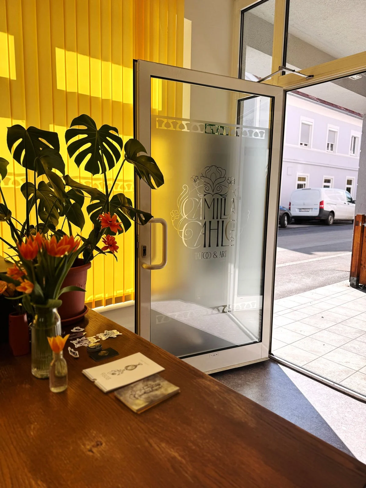 Innenaufnahme eines Geschäfts mit offen stehender Glastür, auf dem Fenster sind Ornamente und der Schriftzug „A MILA CHIC TATO & ART“ zu sehen. Im Vordergrund befinden sich Blumen auf einem Holztisch, mit Postkarten und kleinen Gegenständen. Im Hintergrund ist eine graue Gebäudeansicht draußen sichtbar.