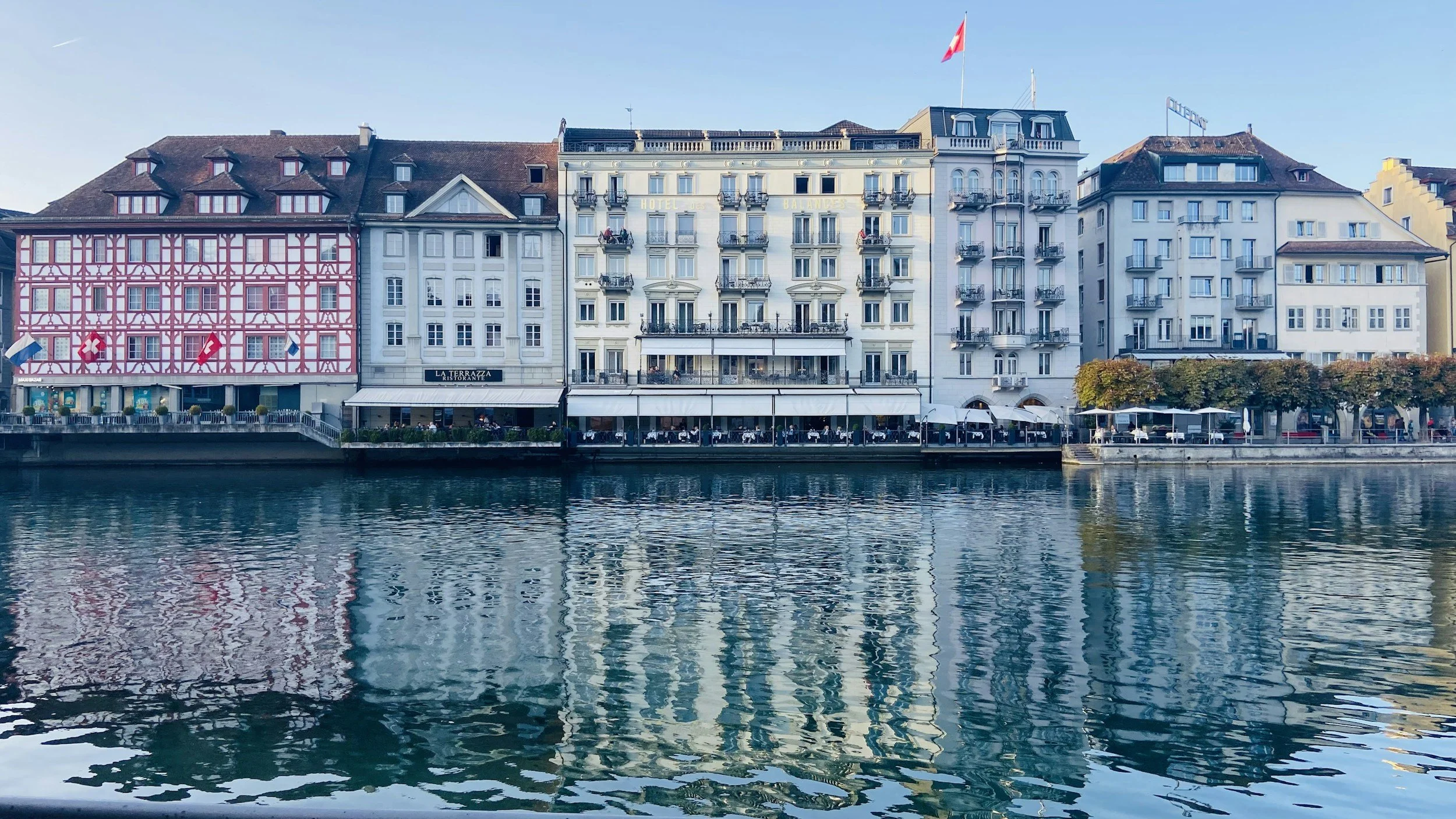 View of waterfront buildings with restaurant and hotel signs, reflecting on water during daytime.