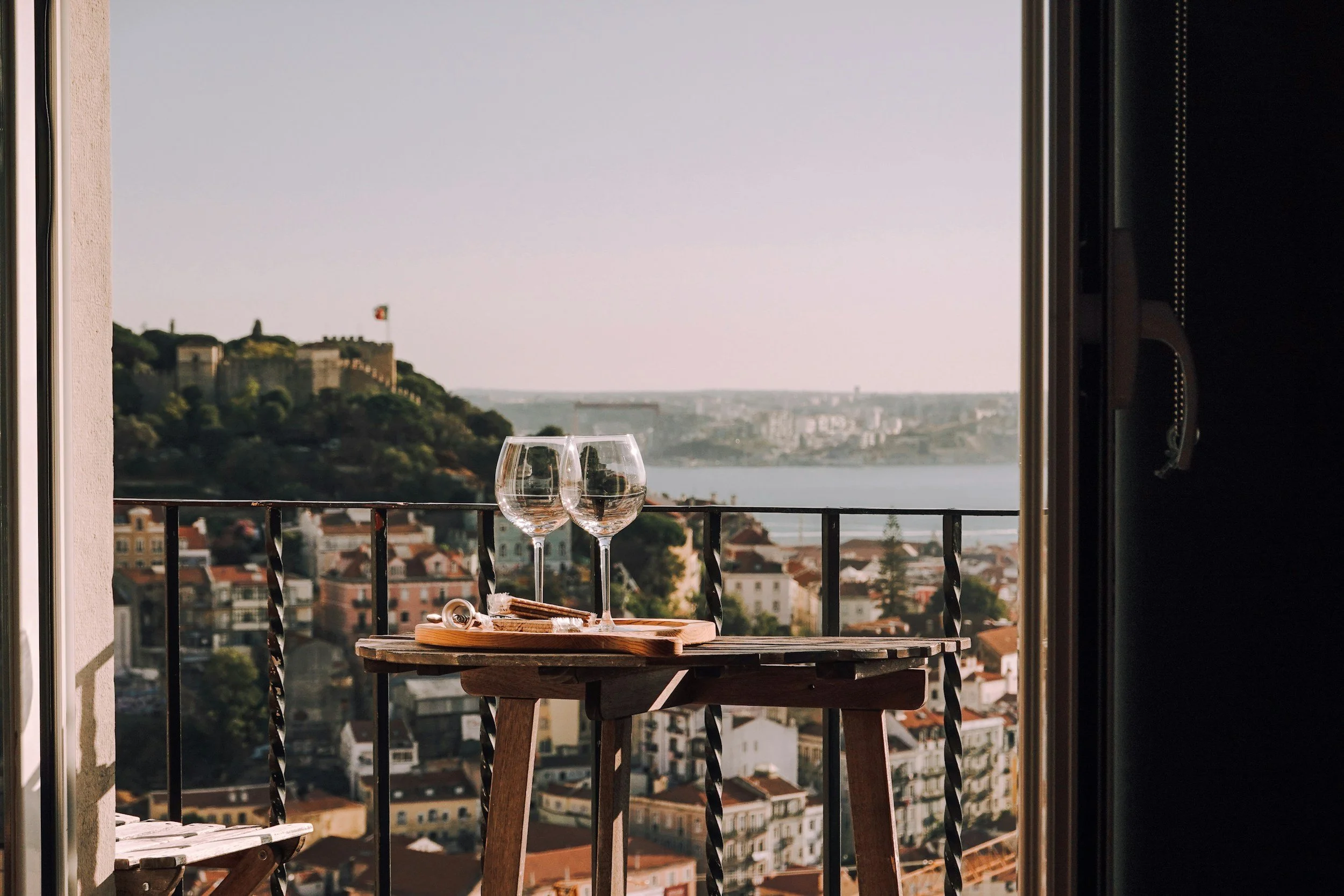 View of a cityscape with a river, hills, and historic buildings from a balcony, with two empty wine glasses on a wooden table.