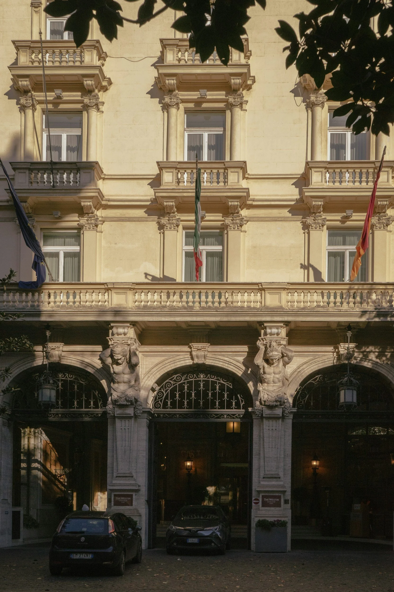 Front view of a historic hotel building with ornate architectural details, statues supporting the upper balconies, three flags flying above, and two cars parked in front.