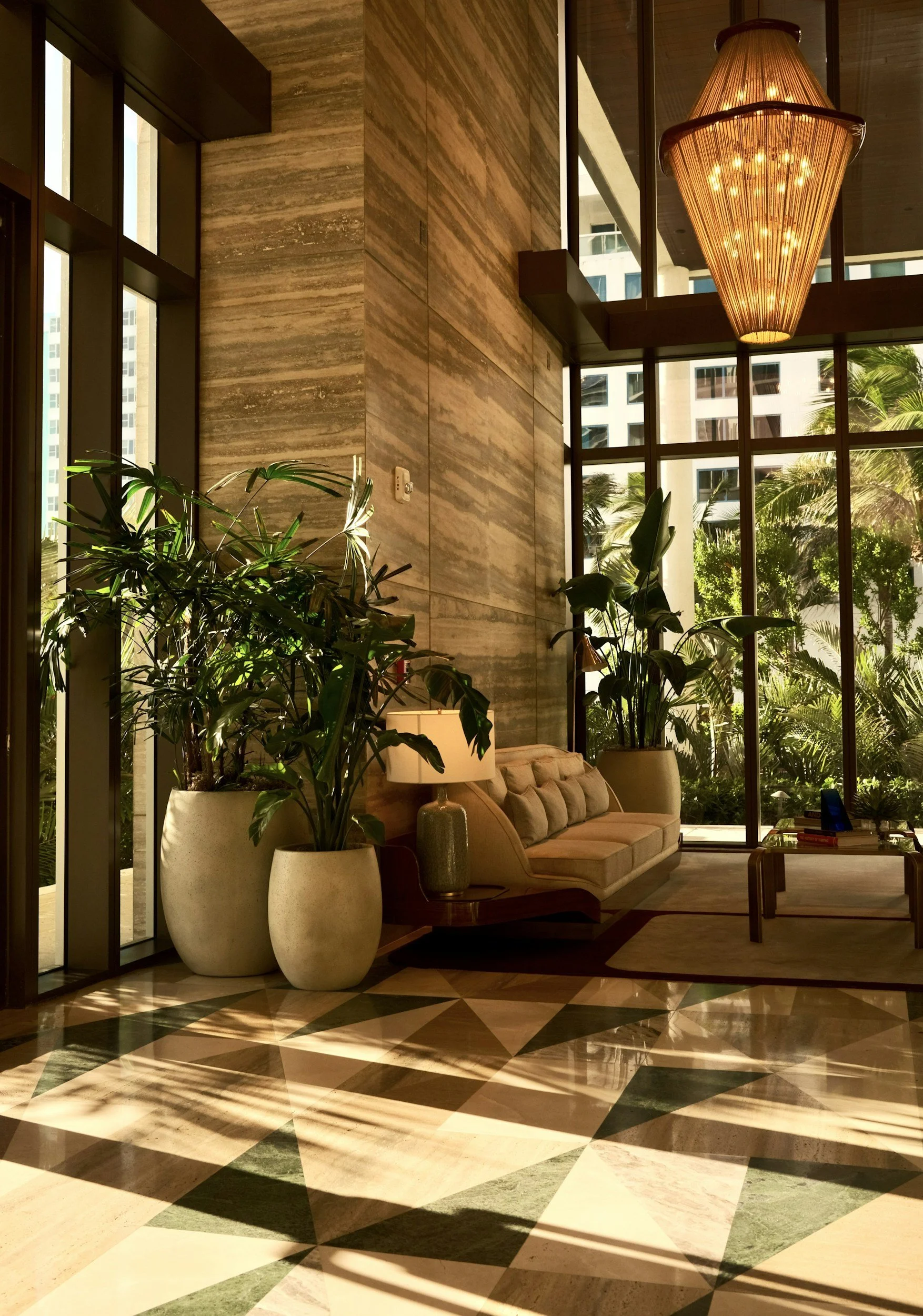 A modern hotel lobby with large windows, tropical plants in white pots, a beige sofa, a large decorative chandelier, and a geometric patterned floor.