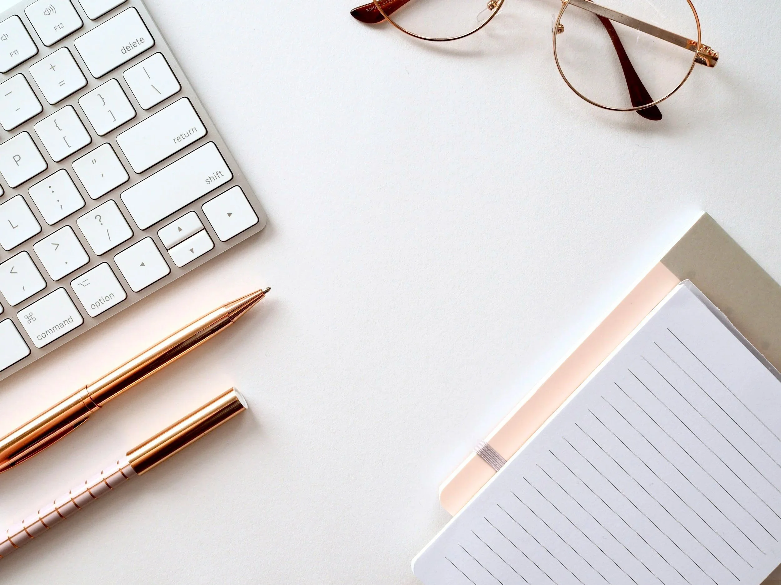 A white desk with a computer keyboard, a pair of round glasses, a rose gold pen and a rose gold pencil, and an open notebook with lined pages.