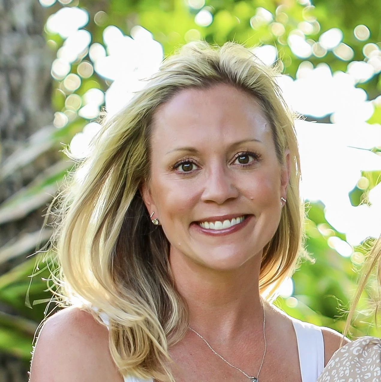 Close-up of a smiling woman with blonde hair, wearing earrings and a necklace, outdoors with green foliage background.