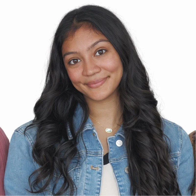 Young woman with long black wavy hair, wearing a denim jacket and a delicate necklace, smiling at the camera.