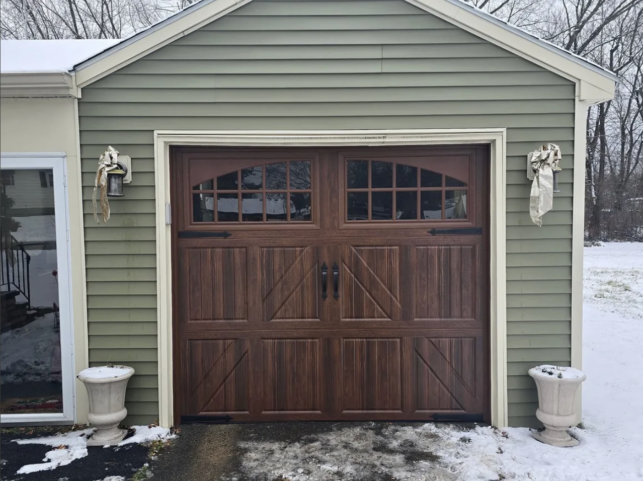 A garage with a wooden door and green siding, snow on the ground, and two large flower pots on each side of the door, one with some snow inside and the other with some debris. Two decorative bows are attached to outdoor light fixtures on either side 